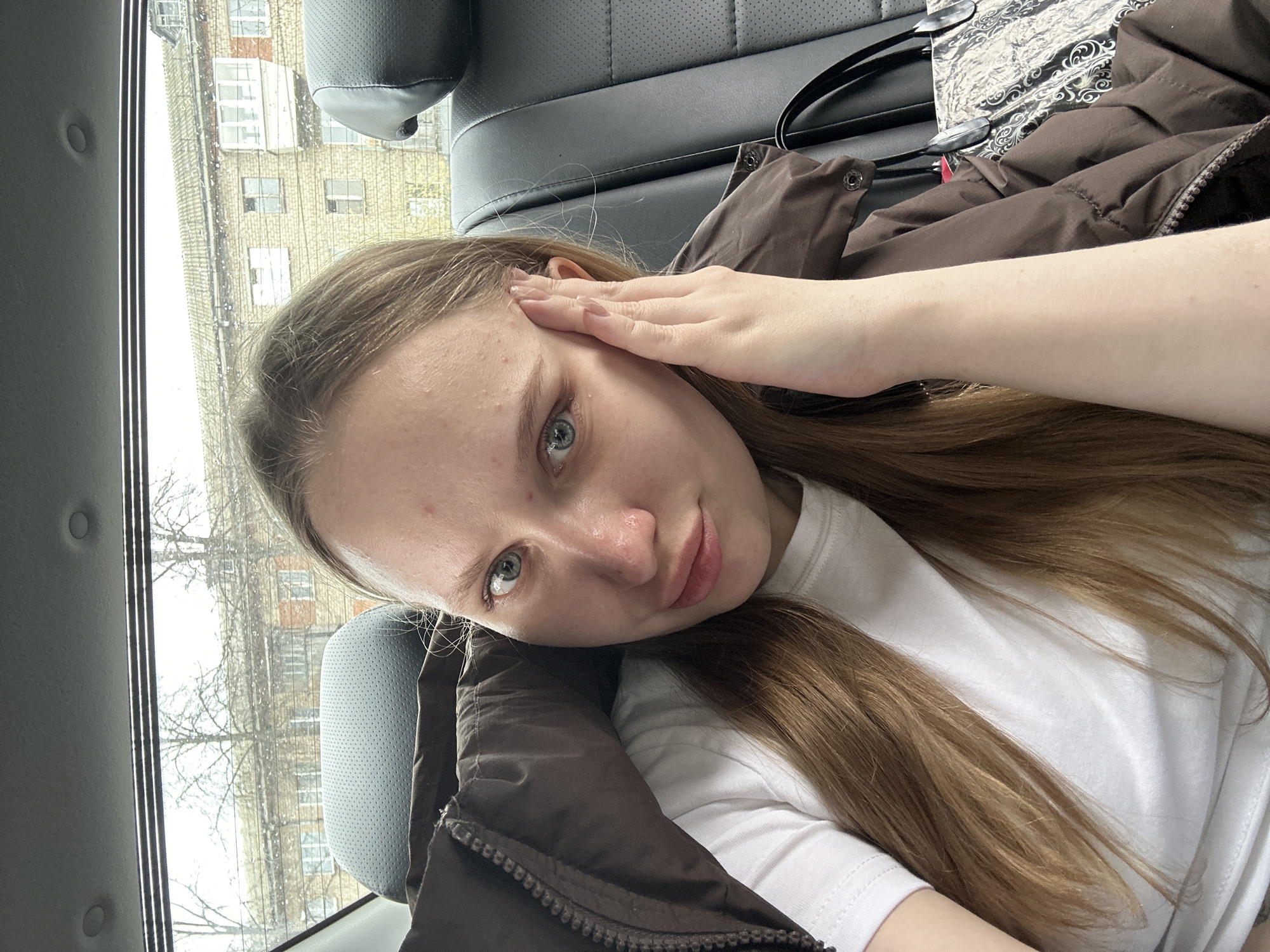 The image captures a candid selfie of a young woman inside a car, presented with a roughly 90-degree clockwise rotation. She has long, light brown hair draped over her chest and shoulders, and is wearing a white t-shirt under a brown puffer jacket, which is partially zipped. Her light blue-grey eyes are directed at the camera, and a gentle smile plays on her lips. Her fair skin has some minor blemishes visible on her forehead. Her left hand is casually placed near her temple, with light-colored fingernails.
The car interior features dark grey or black upholstered seats. Next to her, a black bag with a distinctive patterned flap is visible on the seat.
Outside the car window, an urban or suburban scene unfolds. A multi-story building, appearing to be constructed from brick or stone, with numerous windows, stands prominent. Several bare trees, stripped of their leaves, suggest a late autumn, winter, or early spring setting. The car window itself shows streaks, indicating rain or melting snow. The sky outside appears bright and overcast.
The overall atmosphere is calm and relaxed, captured during daytime. Based on the provided context, the location is Chaikovsky, Russia, with the building's architecture and the seasonal appearance of the trees being consistent with a Russian urban environment during cooler months. There is no visible text in the image.
