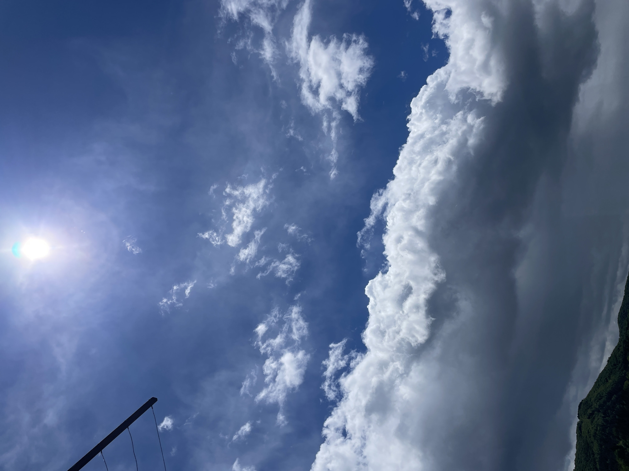 The image captures a view of the sky in Zlatograd, Bulgaria. The sun is visible in the upper left quadrant. Clouds are scattered throughout the sky, ranging from small, fluffy formations to a large, dense cloud bank on the right side of the image. A section of what appears to be a pole with wires attached is located at the lower left corner. The tops of some trees are visible in the lower right corner.