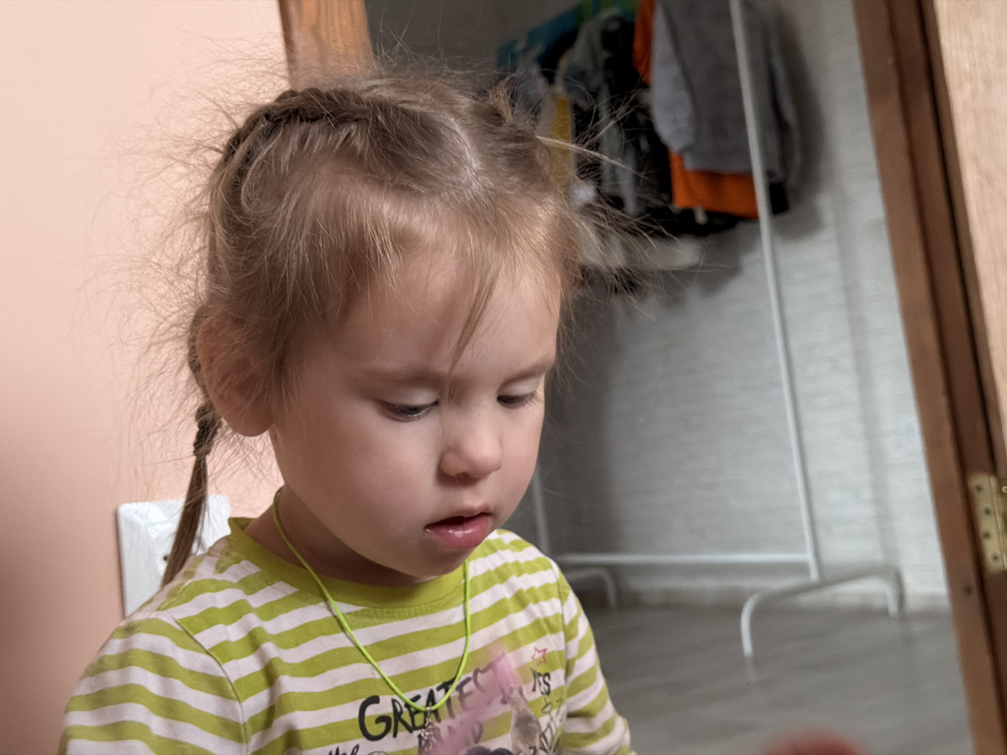 A young child with light brown hair styled in two braids with wisps escaping, is captured indoors. She is wearing a lime green and white striped shirt and a thin green necklace. Her gaze is directed downwards with a slightly open mouth. The background shows a glimpse of a closet with clothes hanging, a white metal clothes rack, and a beige wall. A portion of a wooden doorframe is visible on the right side of the image. The location is Chaikovsky, Russia, based on the available information.