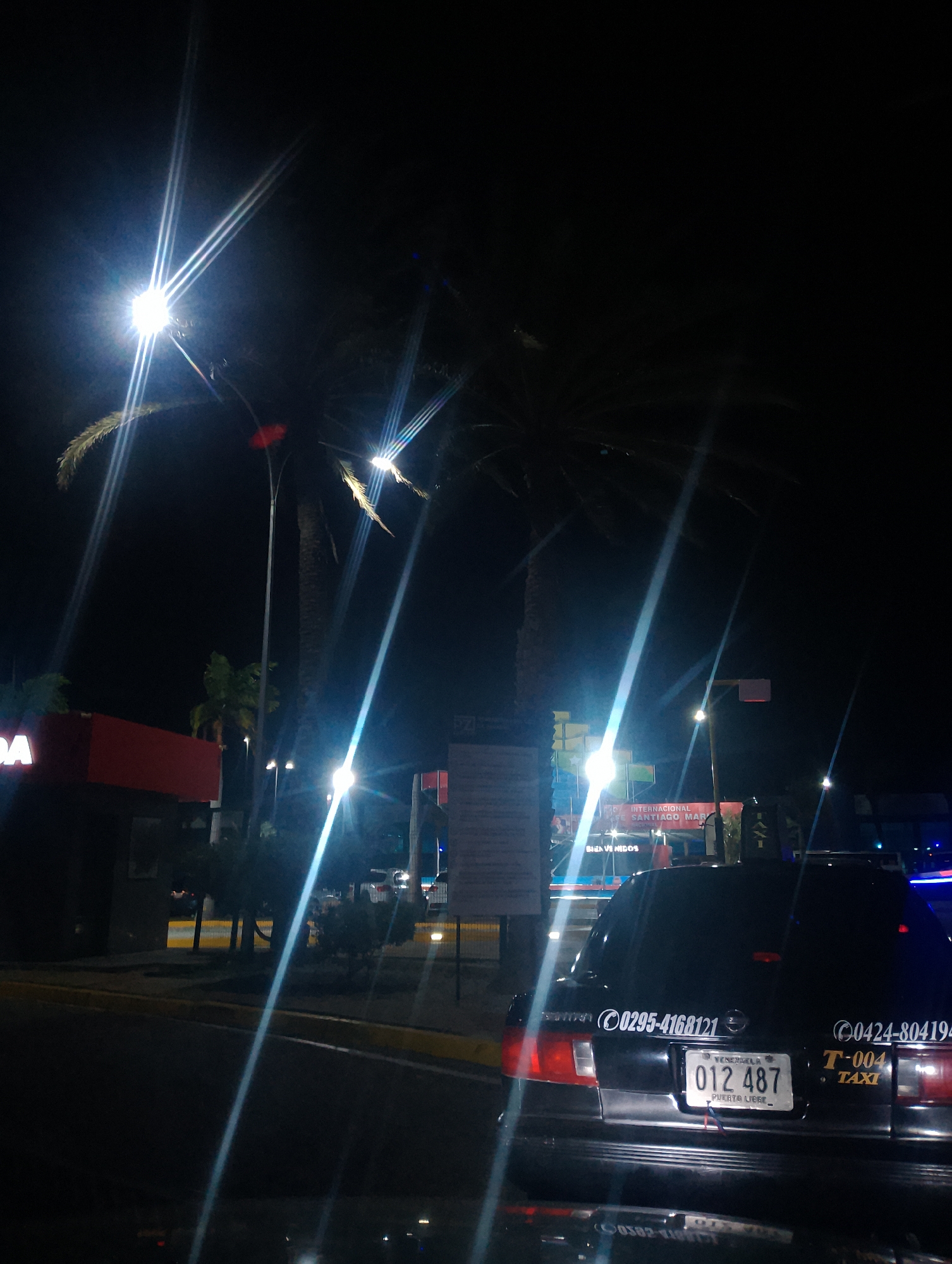 The image captures a night scene in what appears to be an urban area. Prominent are street lights and palm trees, which cast bright glare and lens flare. A black car, identified as a taxi with phone numbers and "TAXI" markings, is in the foreground. Its license plate reads "012 487" and indicates "PUERTO LIBRE" in Venezuela. In the background, there is signage with Spanish text including "INTERNACIONAL DE SANTIAGO MAR" and "BIEN VE NIDOS". There is a red building or structure on the left, and the surroundings suggest a roadside setting at night. Without additional information, the specific location within Cotoperíz cannot be definitively determined.