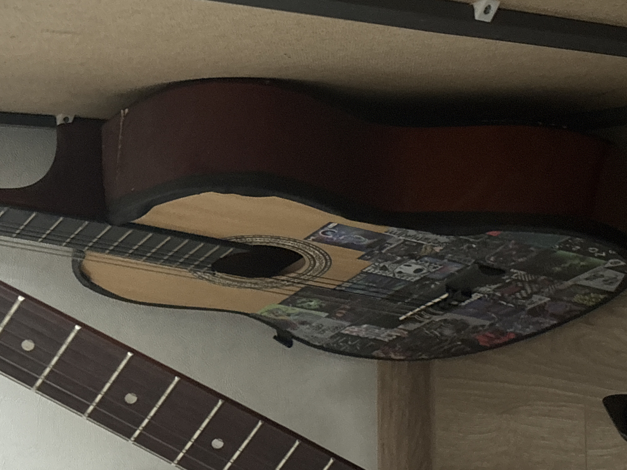 The media file is a close-up, overhead shot of a guitar resting on a shelf. The guitar is a classical acoustic guitar with a light-colored soundboard that is decorated with numerous small, rectangular images, resembling a collage of album covers or photographs. The back and sides of the guitar are a dark reddish-brown color. The soundhole is visible, surrounded by an ornate rosette. The guitar's neck is not fully visible, but a portion of the fretboard with its frets and inlaid position markers can be seen in the foreground, running diagonally across the bottom of the frame.
The setting appears to be indoors, likely a room where the guitar is stored or displayed. The shelf the guitar rests on is visible at the top of the frame, and its surface is a light, neutral color. There are a couple of small white plastic brackets attached to the underside of the shelf, suggesting it is part of a larger shelving unit or furniture. The lighting is somewhat dim, casting shadows and giving the scene a muted feel. There is no text visible in the image. The focus is solely on the guitar and its immediate surroundings.