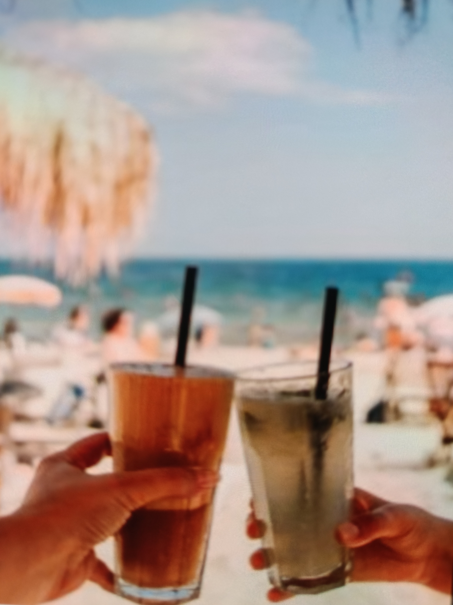 The image is a blurred photograph of two glasses with straws on a beach. The drinks are likely iced coffee or a similar beverage. In the background, there is a beach with people, umbrellas, and the sea. The lighting suggests it is daytime. The photograph is a close-up, focusing on the drinks and the hands holding them. The background is out of focus, indicating a shallow depth of field. Without specific visual cues, the location in Varna, Bulgaria cannot be determined from the image alone.