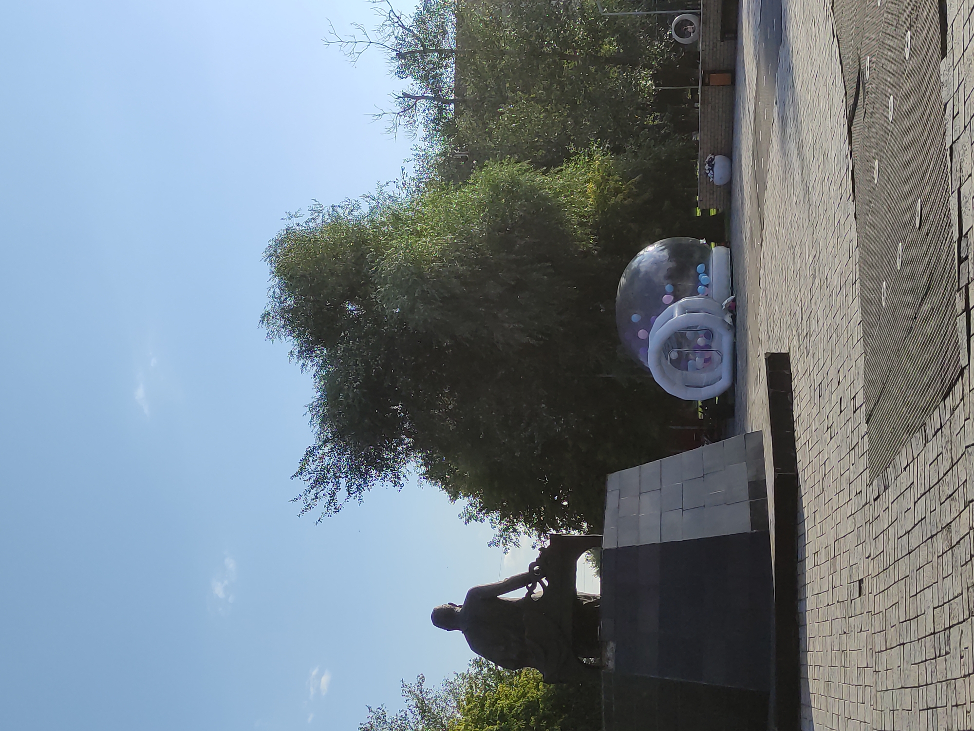 The image shows a wide outdoor plaza in the city of Semey, Kazakhstan. In the foreground, the plaza is paved with light-colored bricks. In the middle ground, there is a large, dark-colored monument with a statue of a seated figure on top. To the right of the monument, there is a clear inflatable bubble with colorful balls inside, and a smaller white inflatable structure next to it. Behind these objects, there are many trees with green foliage. The sky is clear and blue with a few faint clouds. In the far background, there are some buildings and structures, and a person can be seen sitting in a circular opening of a wall.