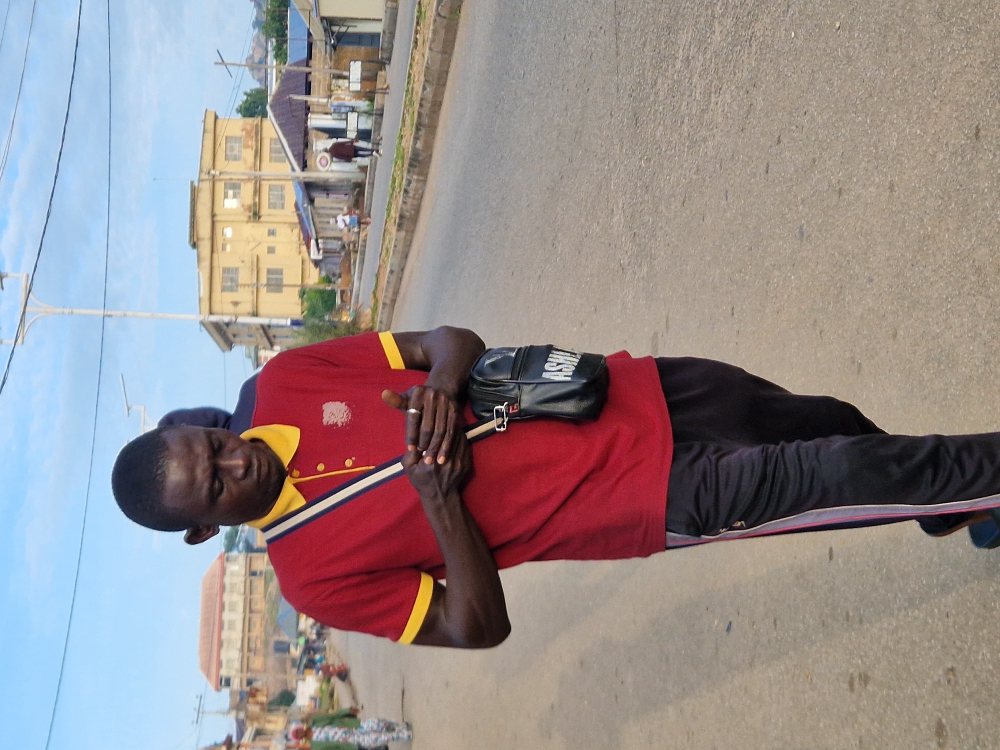 A dark-skinned man, wearing a red polo shirt with yellow trim and black track pants, walks down a paved road in Jos, Nigeria. He is looking at his phone, which he holds in his hands. A black bag with "ASHLEY" written in white letters hangs from his shoulder. In the background, there are buildings, a street light, and other pedestrians. The sky is partly cloudy.