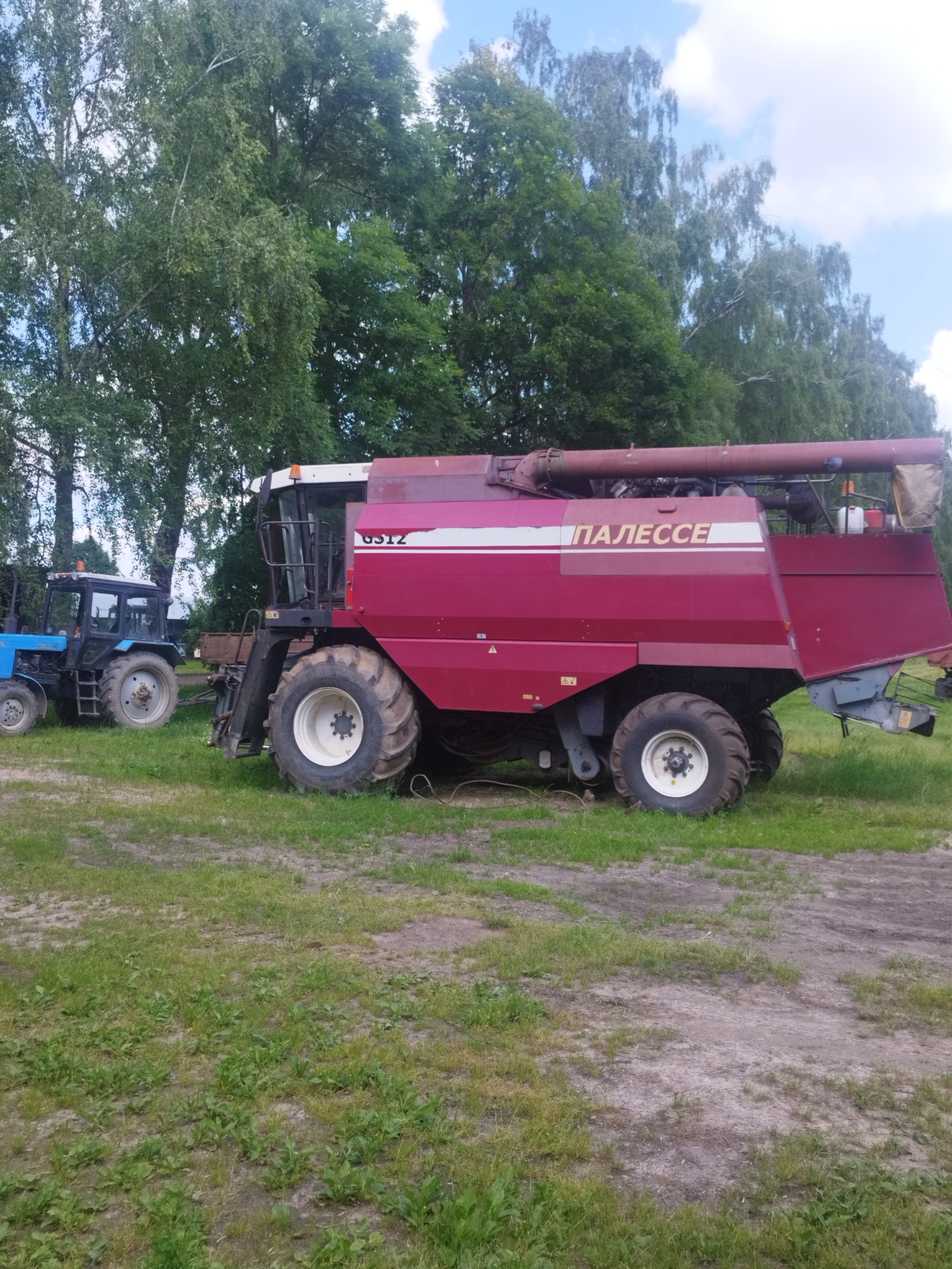 In Залескі сельскі Савет, Belarus, two agricultural vehicles are parked on a grassy area with trees in the background. On the left is a blue Belarus tractor with a cab. To the right of the tractor is a large, maroon combine harvester with the Cyrillic lettering "ПАЛЕССЕ" prominently displayed on its side. The combine also features the model number "GS12". Both vehicles appear to be stationary. The ground is covered with green grass and some dirt patches. The sky above is partly cloudy.