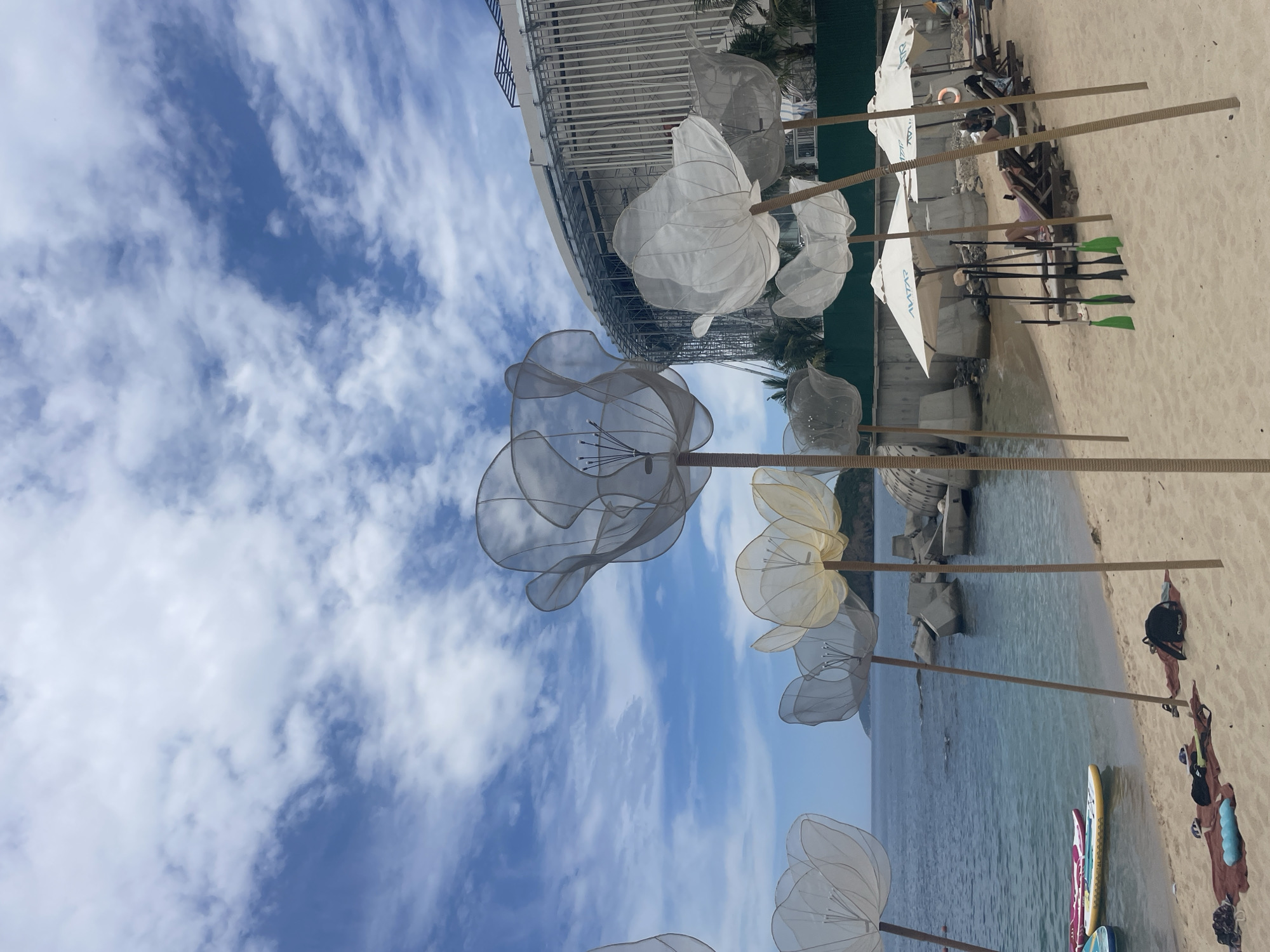 This is a photograph taken at a beach in Nha Trang, Vietnam. The scene is dominated by several large, translucent, flower-shaped sculptures made of white material, placed on wooden poles. In the foreground, a sandy beach is visible with several surfboards resting near the water's edge. Further back, there are lounge chairs, umbrellas, and a few people relaxing. The sea stretches out to the horizon, with a building visible in the background, constructed with a ribbed facade. The sky is blue with scattered white clouds.