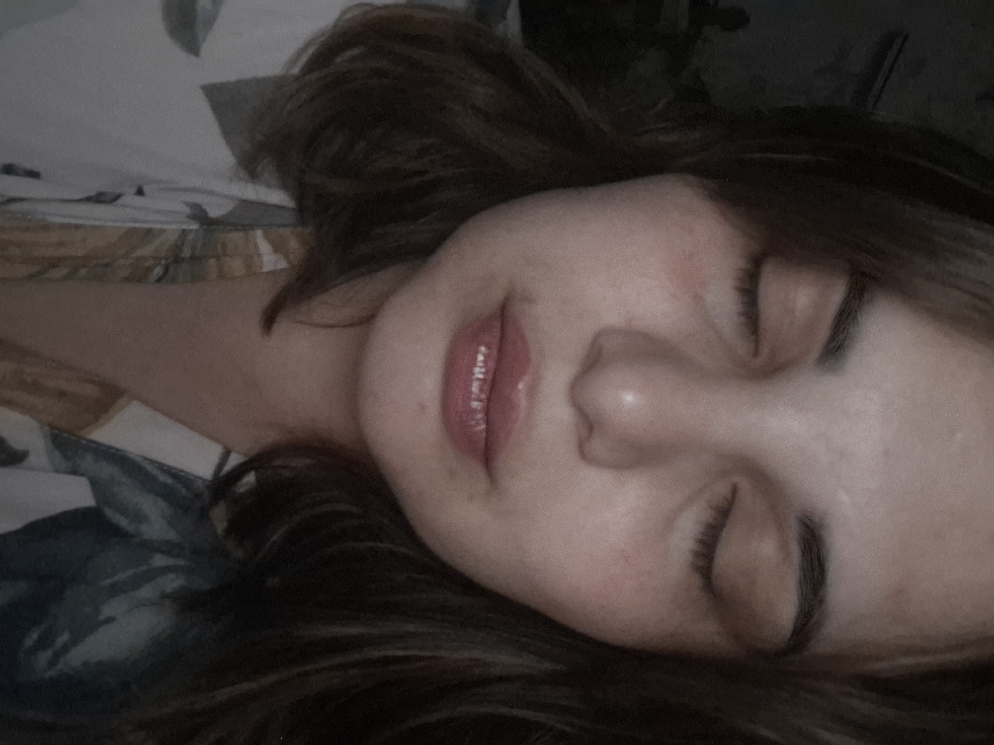 A close-up photograph depicts a woman with fair skin, dark brown hair, and closed eyes, lying on a patterned surface resembling bedding. Her lips are slightly parted, revealing a hint of pink. The background is dimly lit, providing minimal context. There is no information to suggest that the image was taken in the city of Амзя, Russia.