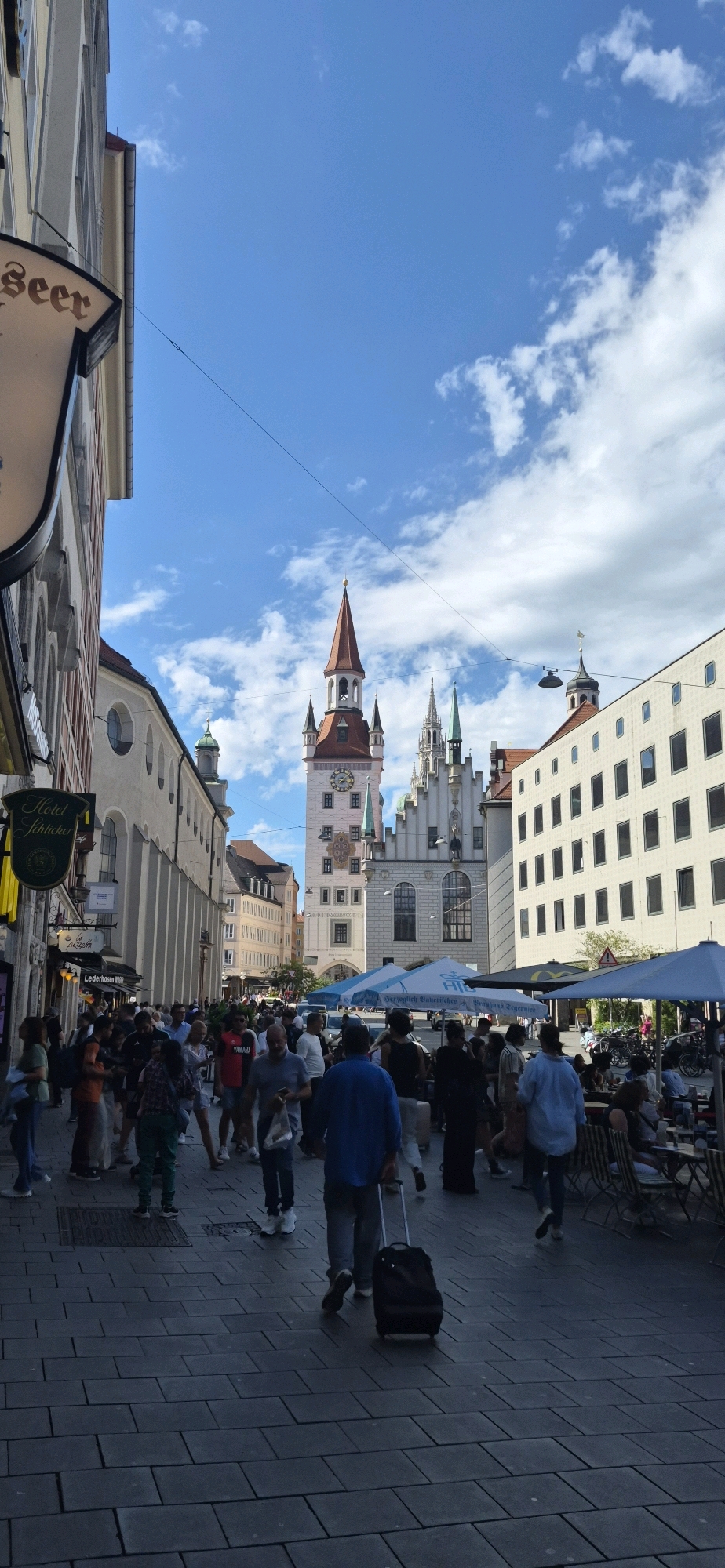 The image captures a bustling street scene in Munich, Germany, likely at the Marienplatz, given the prominent New Town Hall with its distinctive clock tower and spires. The sky is a vibrant blue with scattered white clouds, indicating fair weather.
In the foreground, a paved pedestrian walkway is filled with people. Many are walking in various directions, some with shopping bags, others looking at their phones. A man in a blue shirt is pulling a black wheeled suitcase behind him. To the right, outdoor seating areas for cafes or restaurants are visible, with umbrellas providing shade and people seated at tables.
The buildings lining the street are a mix of architectural styles. To the left, an older building with arched windows is partially visible, along with a sign for "Hotel Schlicker". To the right, a modern building with numerous rectangular windows stands out. In the center, the New Town Hall dominates the skyline with its elaborate facade, clock, and red-tiled roof.
The overall atmosphere suggests a lively tourist area during daytime, with a constant flow of pedestrian traffic and people enjoying the outdoor amenities. The presence of the iconic Munich landmark strongly suggests this is a central part of the city.