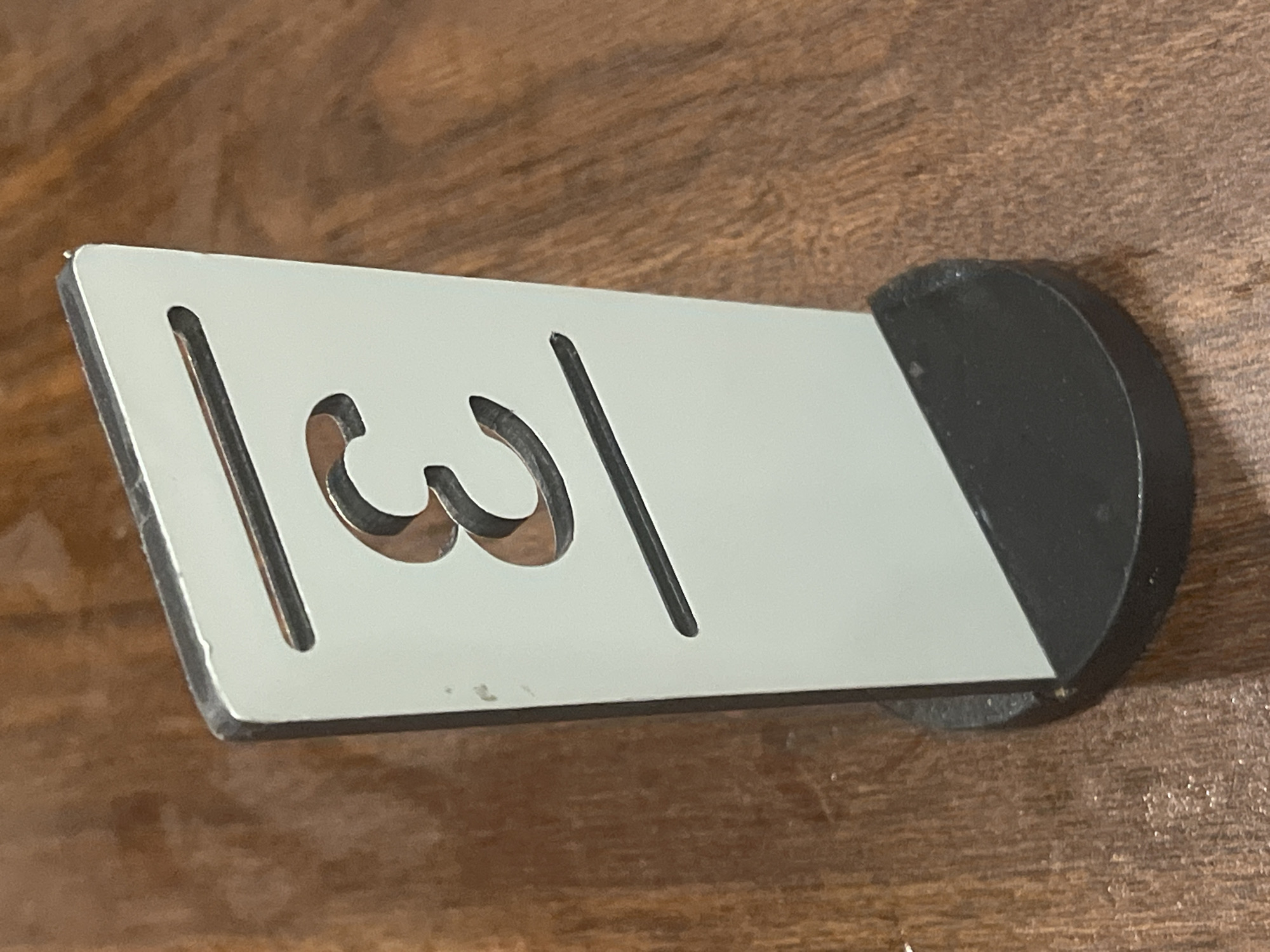 A table number "3" is displayed on a light gray, rectangular sign with a cutout number. The sign has two horizontal slits above and below the number. It stands vertically on a black, circular base, which is placed on a wooden surface with a visible grain pattern. The image does not provide any information about the city of Tanjung Balai Karimun, Indonesia, or any people, actions, or interactions.