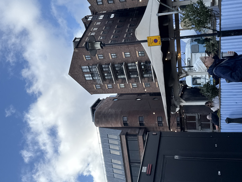 An upward shot captures a brick building with many windows, contrasted against a blue sky filled with fluffy clouds. A smaller, cylindrical tower-like brick structure adjoins the main building. A partially visible black trailer and an outdoor dining area with people present occupy the foreground. A yellow sign, possibly a parking restriction notice, is fixed to a pole.