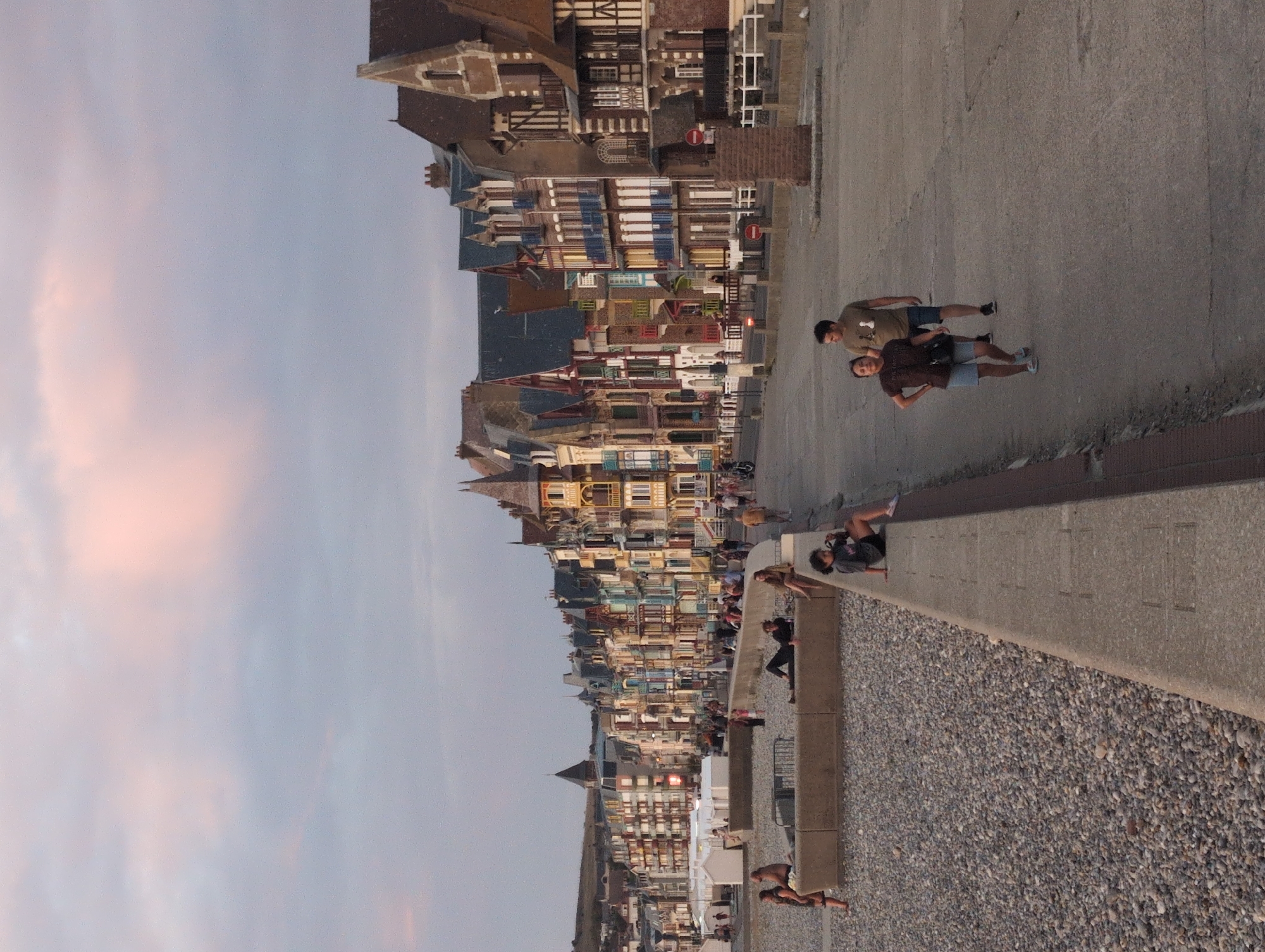This is a medium shot, eye-level, horizontal photograph of the beachfront promenade in Le Tréport, France, at dusk. The sky is a soft, muted pastel pink and grey, with some clouds visible. The buildings lining the promenade are multi-story, brightly colored, and feature varied architectural styles with prominent gables and balconies. Several people are visible. On the beach in the foreground, a woman in a dark top and shorts sits on a concrete barrier, facing left, with her legs crossed and her arms resting on her knees. Further back on the beach, another person is lying down on a similar barrier, and to the far left, a woman in a patterned top and skirt stands with her hands on her hips. On the promenade itself, a couple walks away from the camera towards the right; the man wears a light brown t-shirt and blue shorts, while the woman wears a dark t-shirt and light blue shorts. The promenade is paved and curves to the right, with a low concrete wall separating it from the pebbled beach on the left. In the background, more buildings extend along the coastline, and a gentle hill can be seen in the distance. The overall atmosphere is one of a quiet, relaxed evening by the sea.