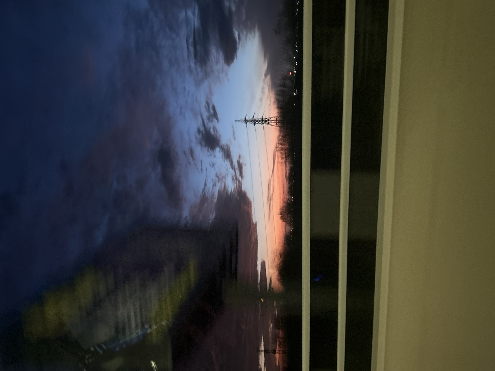The image depicts a power line against a sky transitioning from blue to orange, potentially at sunrise or sunset. The view is likely from inside a building, as suggested by the vertical blinds in the right portion of the image. A city skyline is faintly visible in the background. The reflection of objects is visible on the window. There is no information in the image about the location or the city of Burgas, Bulgaria.