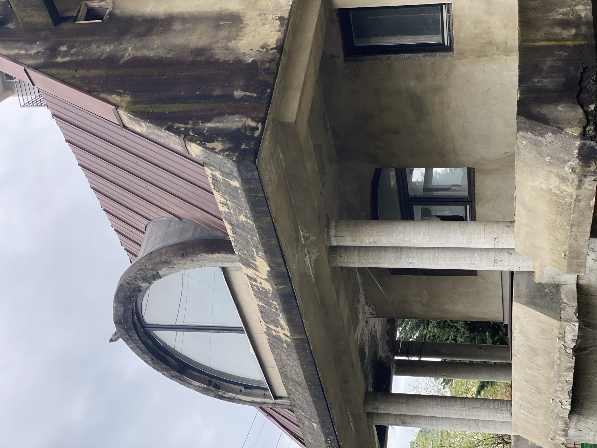 This image depicts a unique architectural structure, possibly a building or a detail of one, in what seems to be a state of age or disrepair. The location is specified as Sochi, Russia, but further information about the exact location within the city is not provided.
The primary subject is a concrete structure exhibiting signs of weathering, with dampness and possible mold growth evident on the concrete and metal roof. The building features distinct architectural elements, including a circular window and a row of cylindrical columns providing support. The windows are covered with glass panels, showing reflections of the outside environment.
The surrounding environment is only partially visible, consisting of trees and a cloudy sky. There are no people present in the image, and the scene is static. There are no notable interactions or actions taking place. The scene has an overall impression of age, and possible abandonment or lack of recent maintenance.