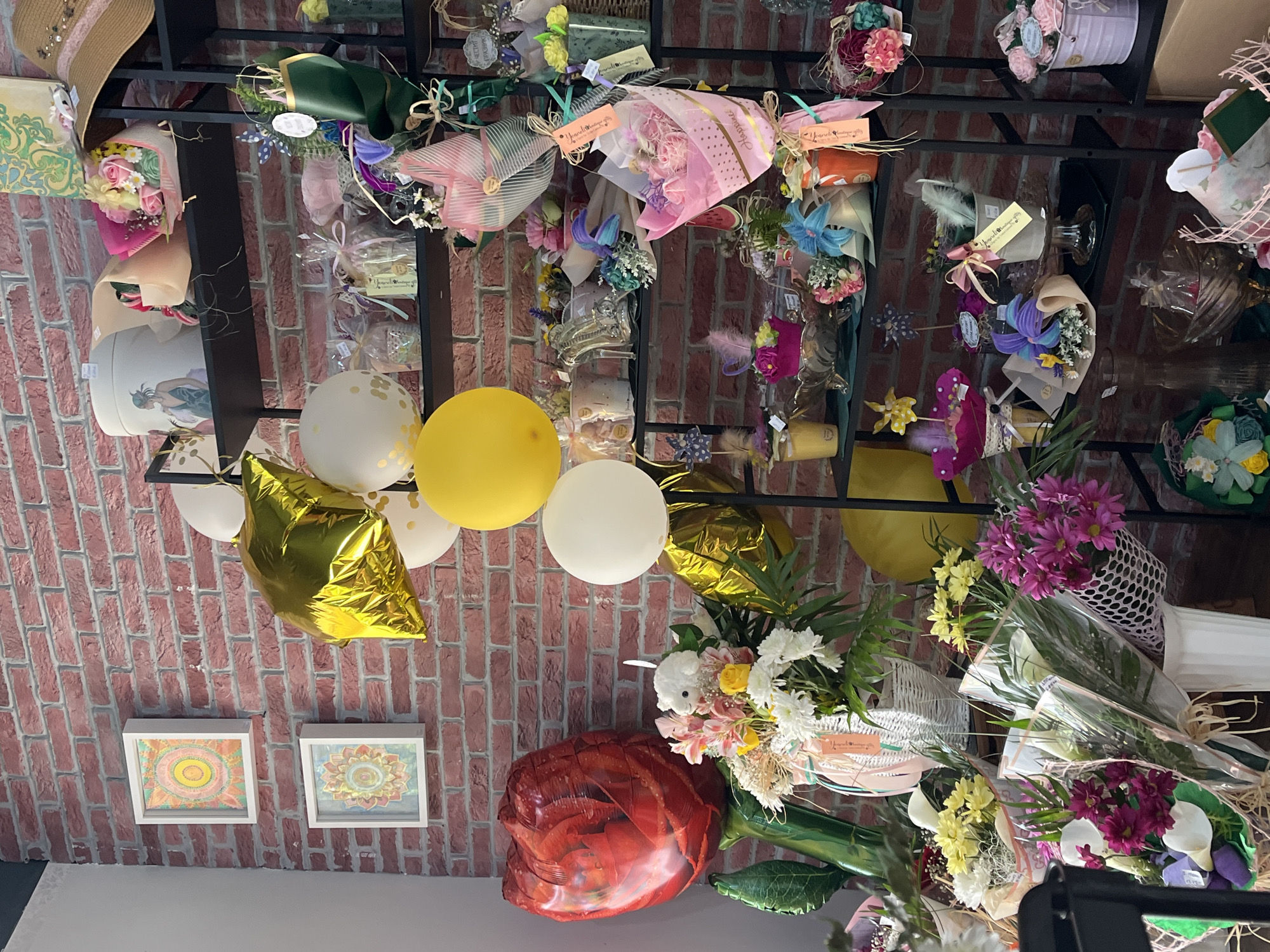 A shop's ceiling is adorned with a variety of items including flower bouquets wrapped in decorative paper, hanging from a metal grid. Balloons of various colors (white, yellow, and gold) are also present. A large red balloon, resembling a flower, is visible. Below, two framed circular images hang on a brick wall. Based on "Yuanzhi Handmade Bouquet" tags in the image, it is likely in the city of Burgas, Bulgaria. The items suggest a flower shop or gift store.