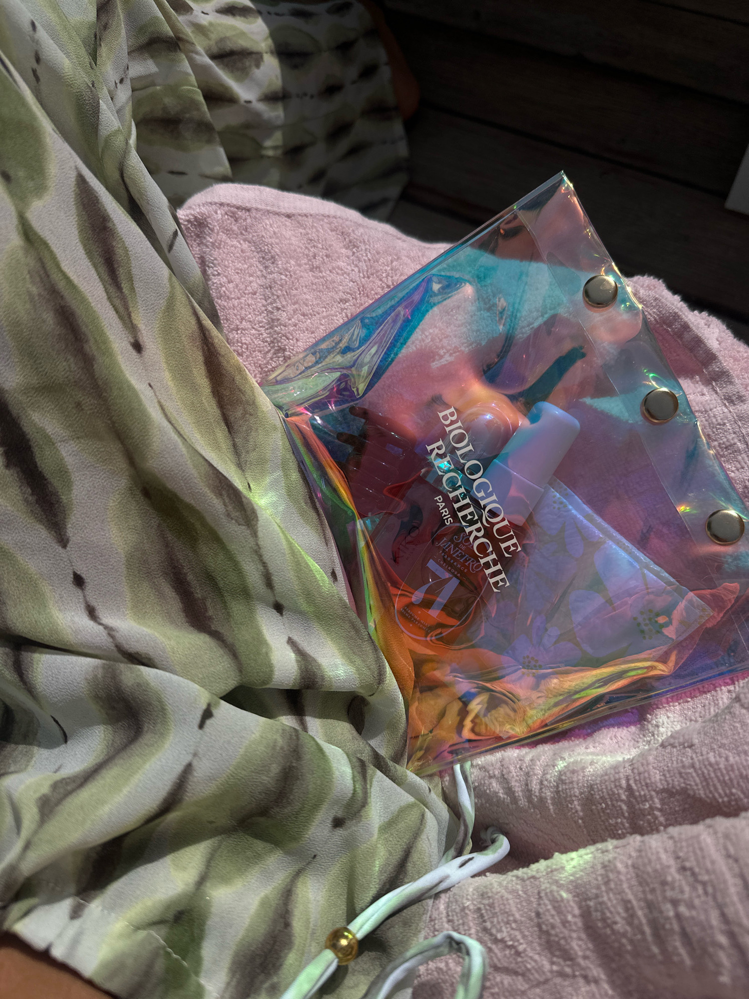 The image shows a close-up of a collection of items resting on a pink textured surface, possibly a towel, with a patterned fabric in shades of green and gray draped over the left side. Dominating the center is a translucent, iridescent pouch, secured with gold-toned snaps. Inside the pouch, several cosmetic products are visible. One product features prominent text reading "BIOLOGIQUE RECHERCHE PARIS." Another item appears to be a small bottle with a light purple cap and a label that includes "SOL DE JANEIRO" and "FRAGRANCE." The overall lighting suggests an outdoor setting, with sunlight casting shadows and highlighting the iridescent qualities of the pouch. The scene is devoid of any discernible urban or natural landmarks that would place it specifically in Pallini Municipal Unit, Greece.