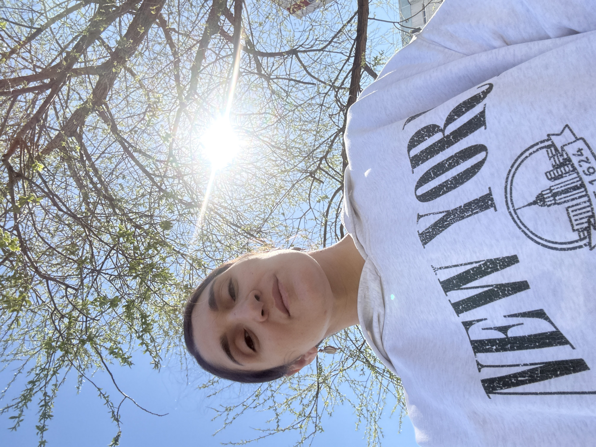 A selfie taken from a low angle features a person wearing a light grey sweatshirt with "NEW YORK" printed on it. The person's face is centered in the lower portion of the frame. Above, tree branches with sparse green leaves are silhouetted against a bright blue sky with a sun flare near the top. The location is unknown, as there are no distinguishing features to indicate it is in Tobolsk, Russia.