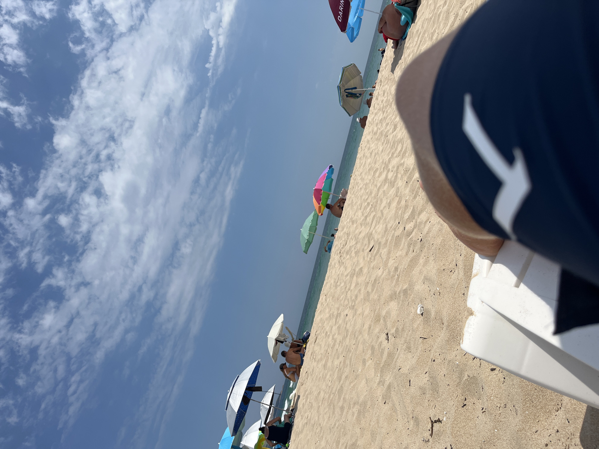 The image depicts a crowded beach scene in Primorsko, Bulgaria, on a sunny day. The foreground is dominated by a person lying on a lounge chair, with their dark blue swim trunks and a white logo visible. The sand is golden and speckled with small debris. In the mid-ground, a slope of sand leads down to the turquoise sea, where several people are visible relaxing under various colorful beach umbrellas. Umbrellas of different colors, including blue, white, green, and a rainbow-striped one, dot the sandy expanse. Further down the beach, more individuals are seen sitting or lying on the sand, some partially obscured by umbrellas. The sky is a clear blue with wispy white clouds, indicating good weather. The overall impression is one of a popular tourist destination with a vibrant atmosphere.