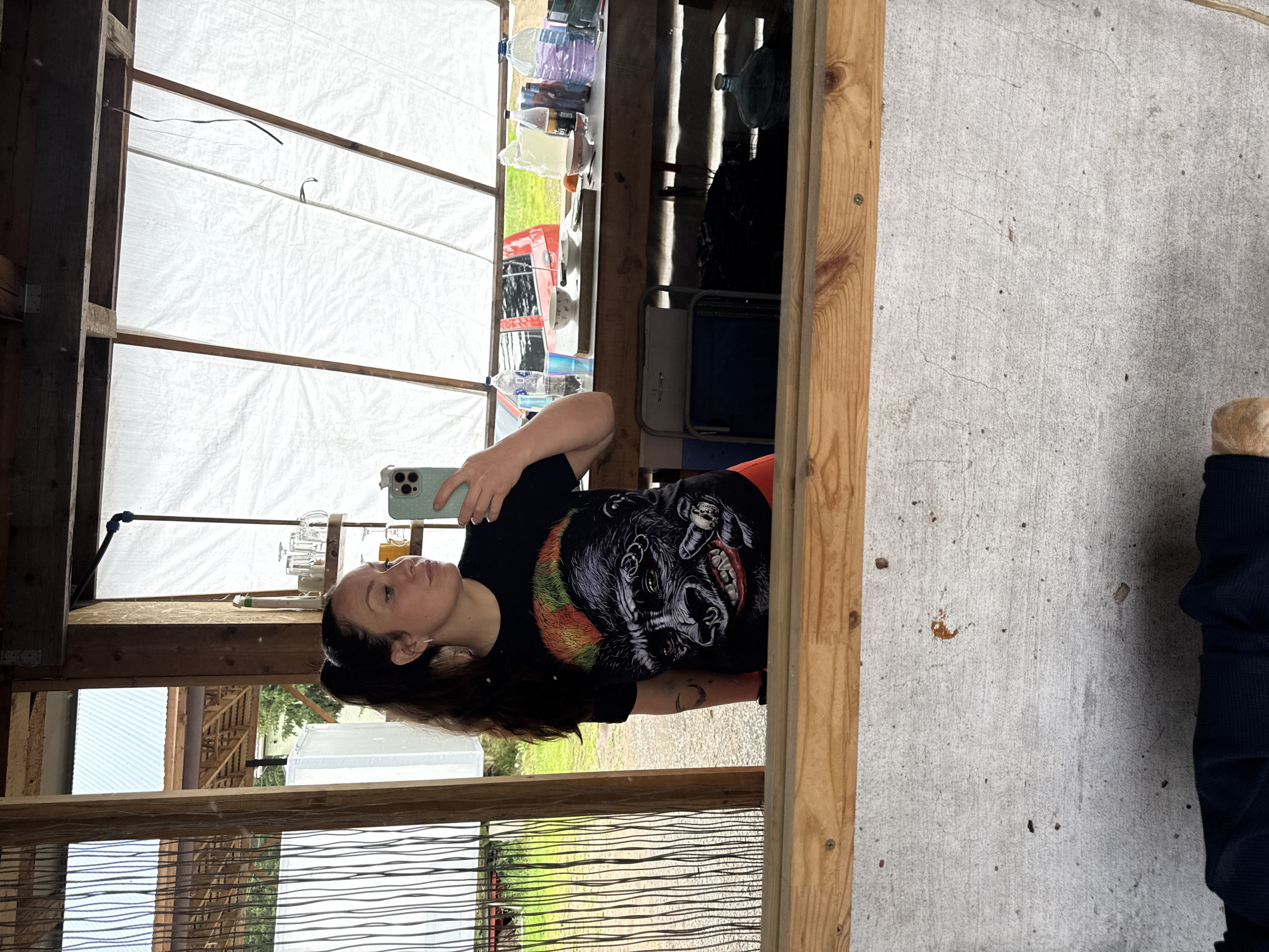 A young woman with a high ponytail, wearing a black t-shirt with a graphic of a gorilla's face and orange shorts, stands in front of a mirror. She is holding a smartphone in her right hand, taking a selfie. The background appears to be an open-air structure with wooden beams and translucent white sheeting. A table behind her is laden with bottles, glasses, bowls, and other items. Outside the structure, green grass and trees are visible, suggesting a semi-rural or outdoor setting. The presence of a car in the background hints at a location within a populated area. No specific landmarks in Chaikovsky, Russia, are identifiable from the image.