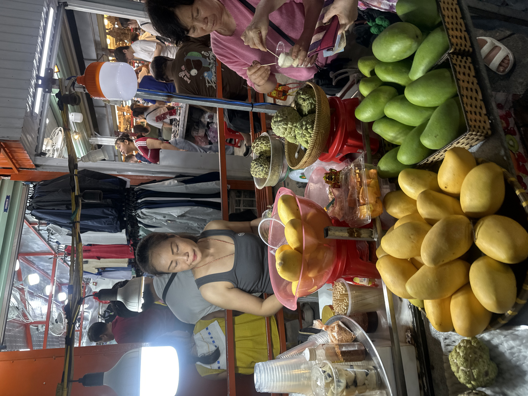 The image depicts a bustling scene in what appears to be a market stall in Đà Nẵng, Vietnam. A vendor, a woman in a grey tank top, is behind a stall selling fresh fruits. Various fruits are visible, including green mangoes, ripe yellow mangoes, and custard apples. Other market-goers are present, including a woman in a pink shirt sampling food with a toothpick. Clothing racks and other merchandise are visible in the background, indicating a diverse market. The lighting is bright, a mixture of natural and artificial light from overhead bulbs. The focus is on the produce and the vendor in the foreground.