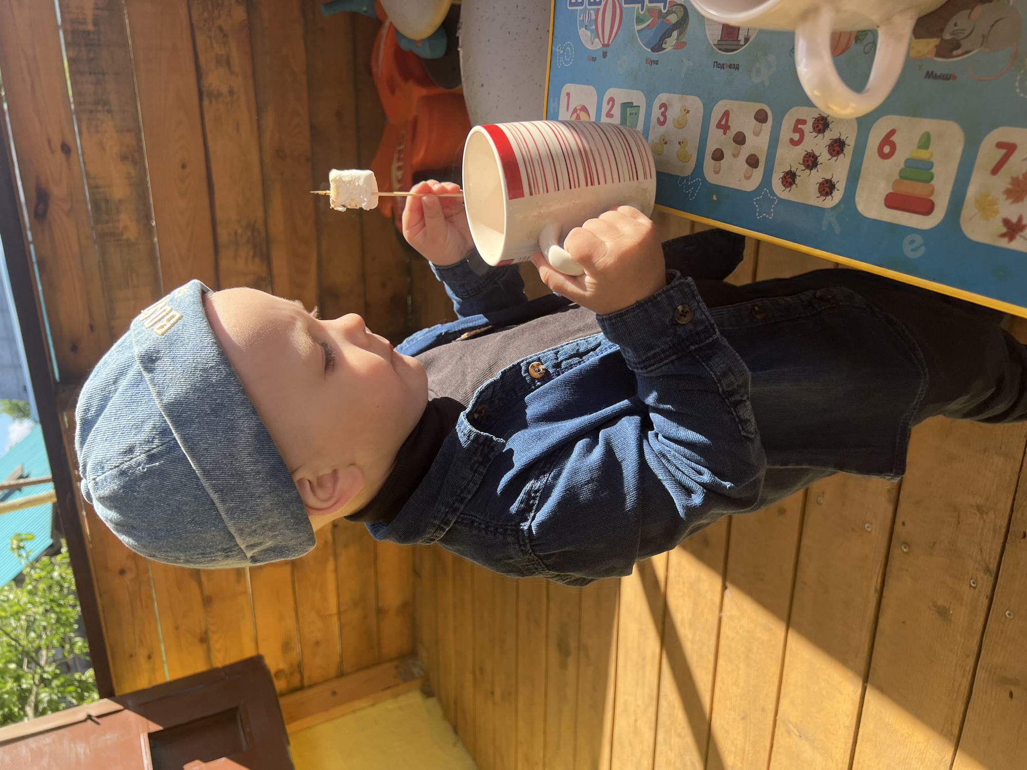 A young child, possibly a toddler, stands outdoors on a wooden deck or porch. The location is possibly in Noviy, Russia based on contextual details that are not explicitly visible in the image, the child is wearing a denim hat and jacket. The child holds a white mug with red stripes in one hand, and a stick with a marshmallow impaled on it in the other. A children's educational mat with numbers and pictures is visible nearby. The background includes wooden siding and glimpses of foliage. The scene is brightly lit with natural sunlight.