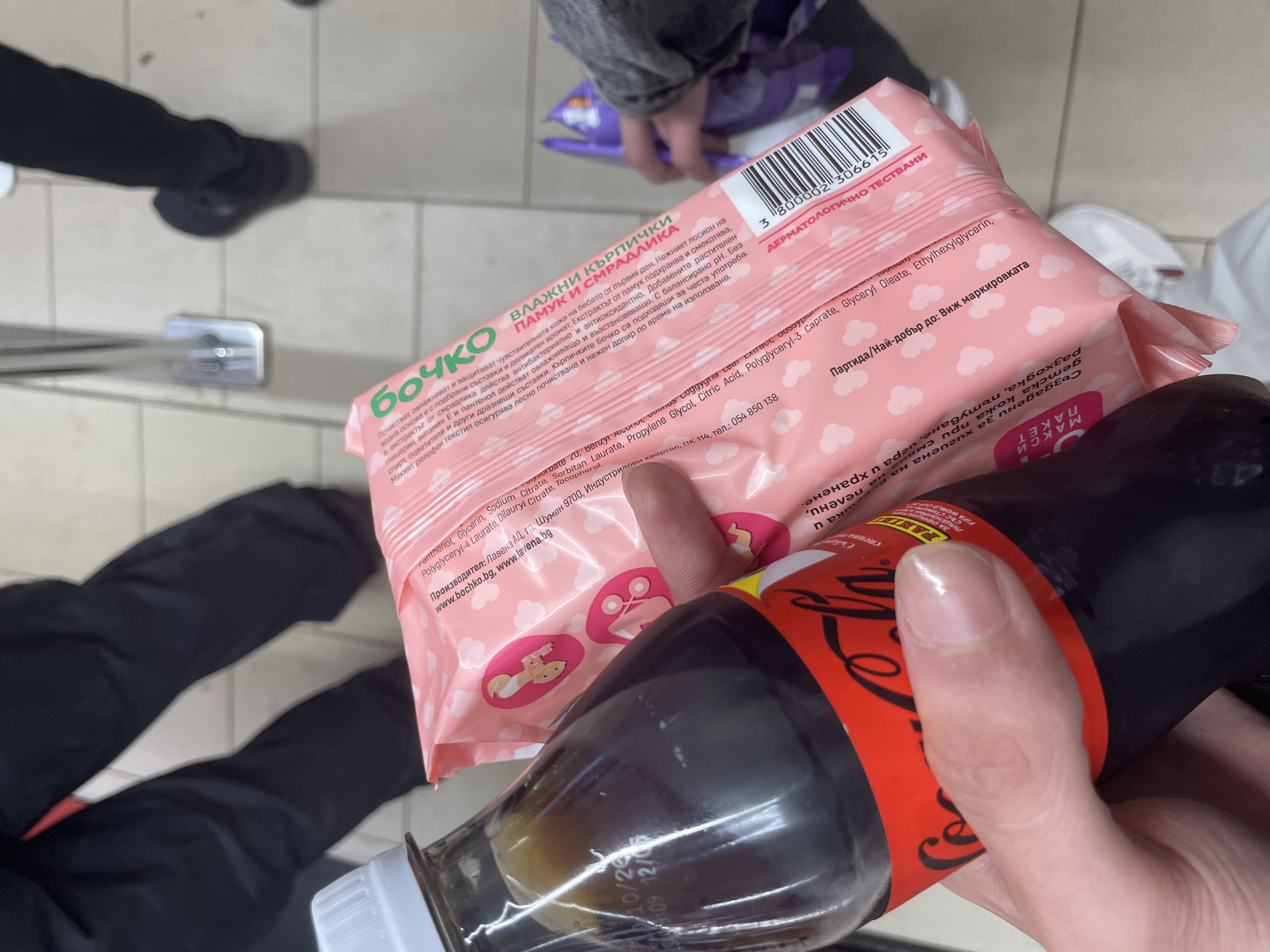 An overhead shot captures a person holding two items in what appears to be a supermarket or convenience store. The setting features a tiled floor with light-colored grout, suggesting an indoor commercial space. In the foreground, a person's hands are visible, holding a pink package of "БОЧКО" (Bochko) brand wet wipes and a dark-colored bottle, likely a Coca-Cola product, recognizable by its distinctive red label and white script. The bottle also displays "PANINI FIFA WORLD CUP" and "Със стикери" (With stickers), indicating a promotional tie-in.
In the background, other shoppers' legs and feet are visible, one pair walking away to the right, and another pair visible to the far left. A purple item, possibly another package, rests on the floor in the mid-ground. A silver metal bar, potentially part of shelving or a display unit, is also visible.
The wet wipes package is predominantly pink with white cloud patterns and illustrations of babies. Extensive Bulgarian text is visible, indicating the product's origin and market, consistent with the provided location context of Ihtiman, Bulgaria. Key phrases include "ВЛАЖНИ КЪРПИЧКИ" (Wet Wipes), "ПАМУК И СМРАДЛИКА" (Cotton and White Horehound), and descriptions of the product's gentle properties for sensitive baby skin, being "ДЕРМАТОЛОГИЧНО ТЕСТВАНИ" (Dermatologically Tested) and free from alcohol and dyes. Manufacturer details for "Лавена АД" in Shumen, Bulgaria, and website addresses are also present. The bottom right of the package displays upside-down text detailing its use for "хигиена на детска кожа при смяна на пелени, разходка, пътуване, игра и хранене." (hygiene of baby skin during diaper changes, walks, travel, play and feeding). A barcode and a partially visible date "10/26 12/05" are seen on the Coca-Cola bottle.