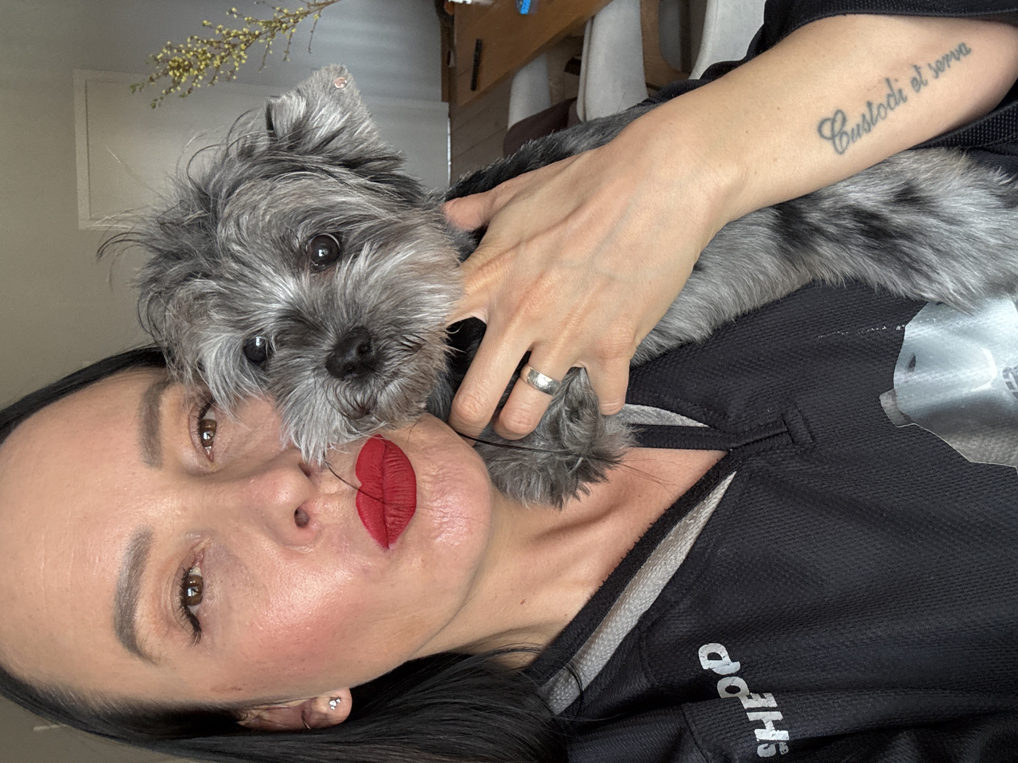 The image is a close-up, selfie-style portrait of an adult woman and a small dog, rotated clockwise. The setting is indoors, with a light-colored wall, a hint of a white frame or doorway, and a decorative plant branch with small yellow clusters visible in the background, suggesting a home environment in Chaikovsky, Russia.
The woman has dark hair and pronounced red lipstick. Her brown eyes look directly at the camera, and her expression is somewhat serious or pouting. She has ear piercings and wears a silver-toned ring on her left ring finger. A tattoo on her right forearm, which is resting on the dog, reads "Custodi et serva" in script. She is wearing a black, textured shirt with grey accents, on which partially inverted text that appears to be "SHQPD" or similar is visible.
Cradled close to her face is a small, scruffy-haired dog, predominantly grey with some darker fur. The dog, likely a terrier mix, has dark, round eyes and a black nose, and also looks attentively at the camera. The woman's hand is gently placed on the dog's back. The activity captured is a shared moment, likely a selfie, highlighting the bond between the woman and her pet. The lighting appears to be soft, natural indoor light.