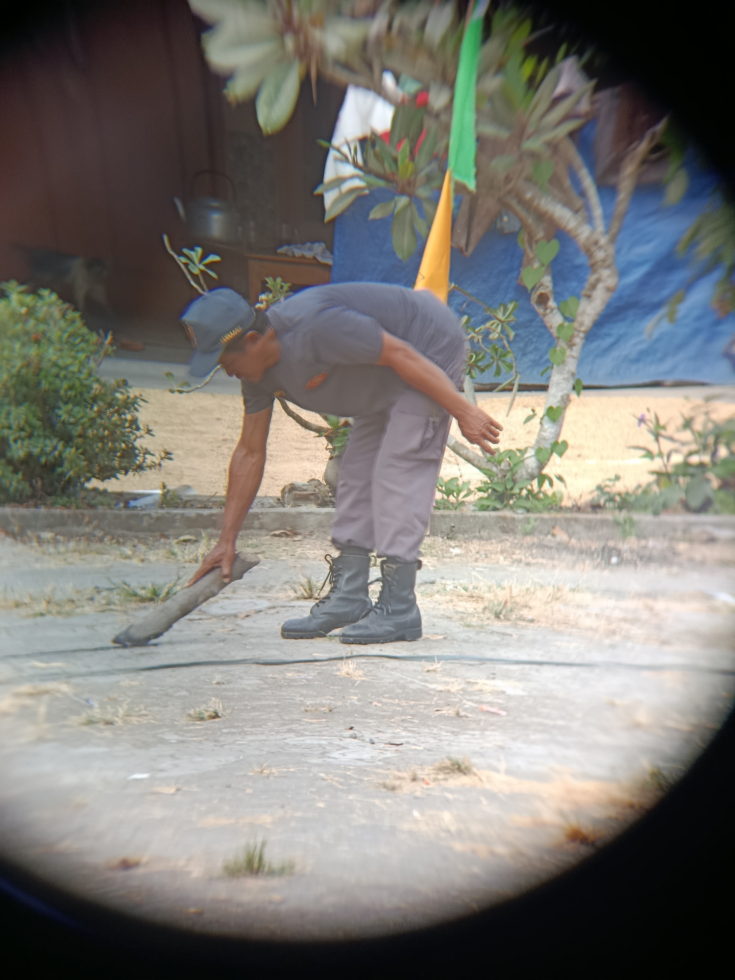 In Sarwadadi, Indonesia, a man wearing a dark t-shirt, grey pants, a baseball cap, and black boots is seen bent over, using a stick to move a piece of debris on a paved ground. He is outdoors, with a brown structure and a tree with green and yellow flags in the background. The scene appears to be in a somewhat overgrown area, with dry grass and plants visible.