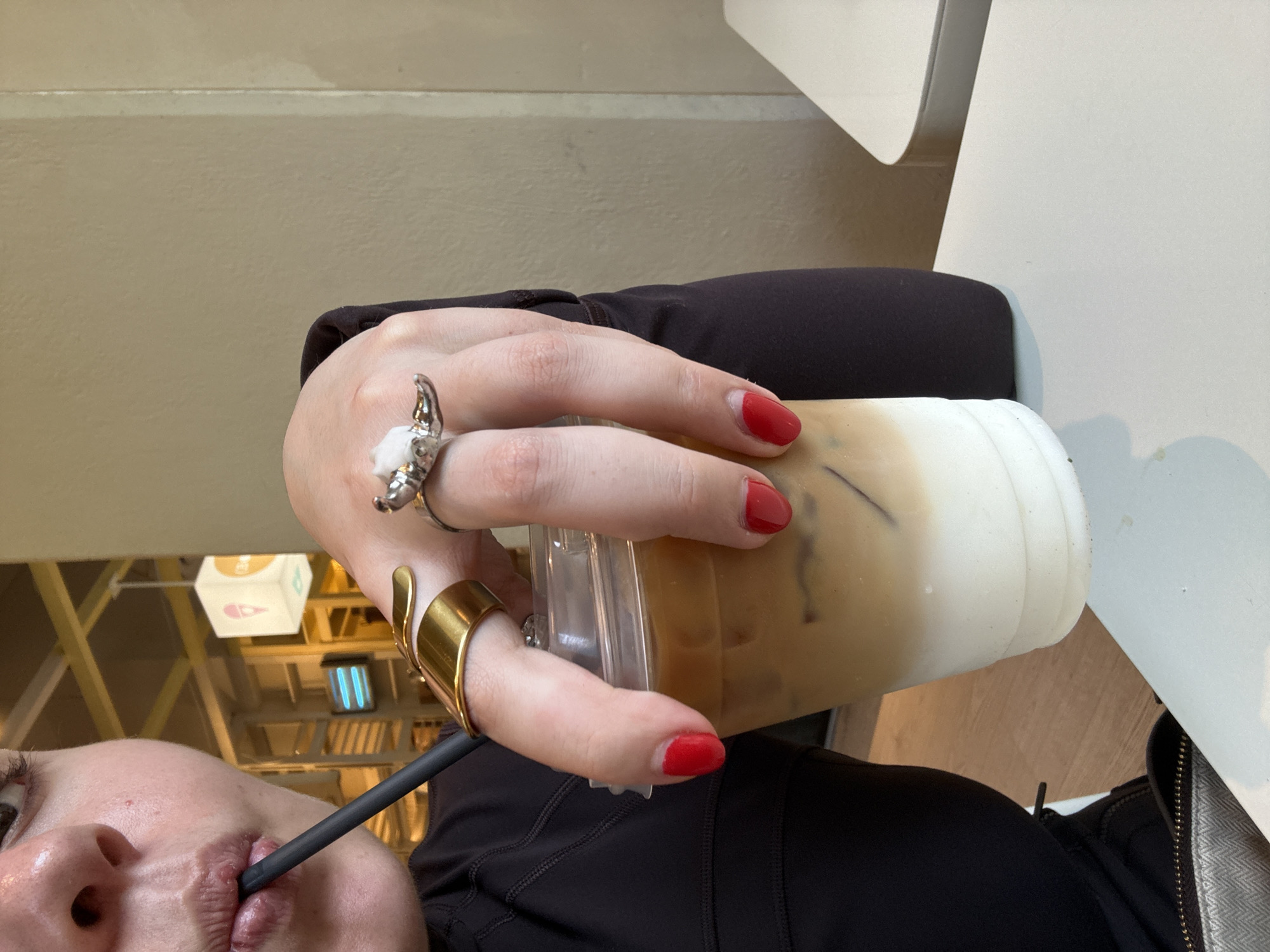 A person is holding an iced coffee in a plastic cup with a straw, with their lips visible on the straw. They are wearing two rings on their left hand: one silver with a white stone and one gold. The fingernails on their left hand are painted red. They are wearing a black shirt. The photo appears to have been taken indoors at a restaurant or café, with a hanging light fixture visible in the background. While the exact location within San Pedro Garza García, Mexico, is not discernible from the image, the presence of the hanging light and the style of the restaurant suggest it is a modern café or dining establishment within the city. A white table is partly visible on the right of the image, and a chair is also partly visible.