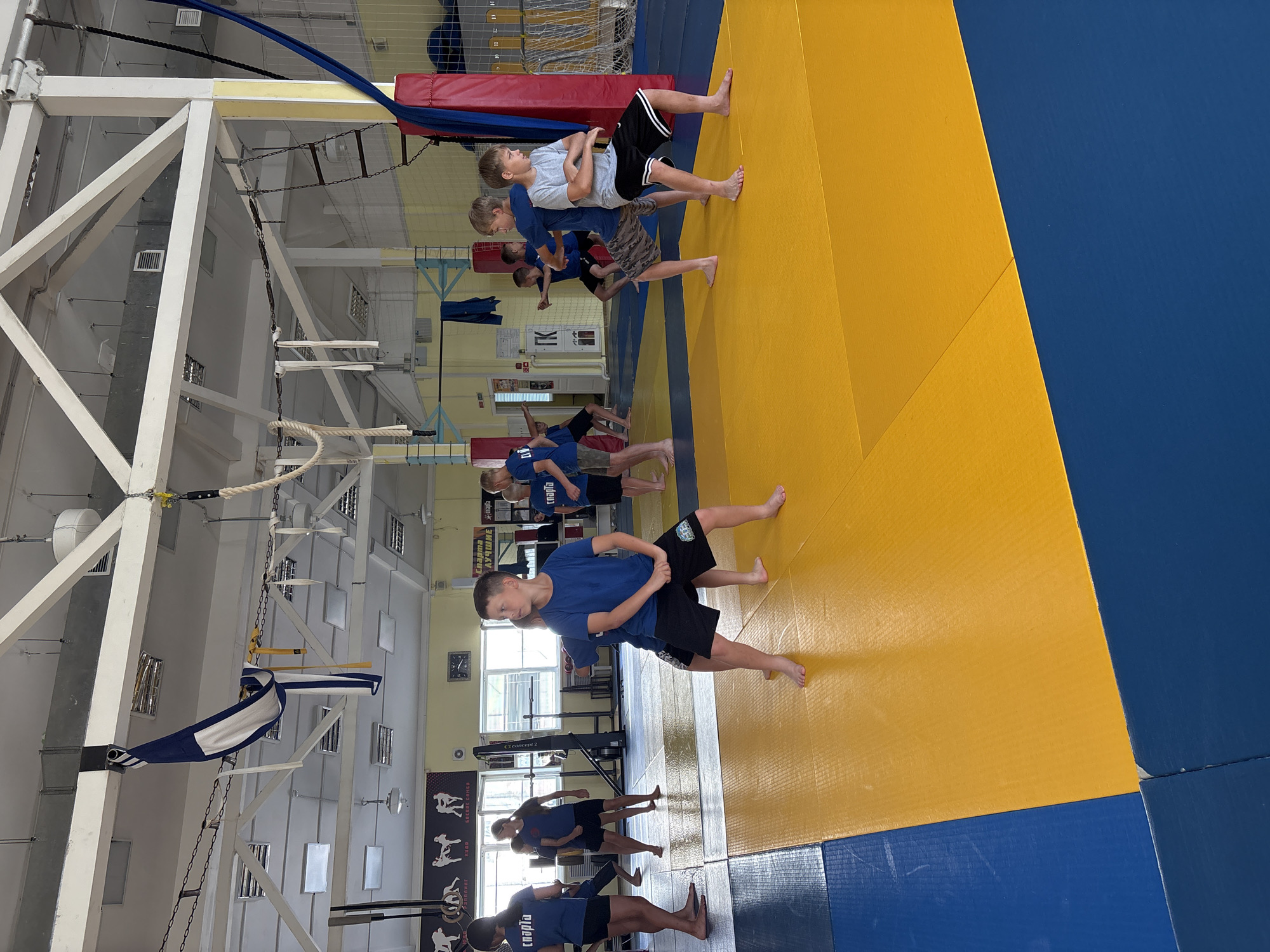 The image depicts an indoor gymnasium with a blue and yellow mat on the floor, typical for martial arts training. Several young boys are present, dressed in athletic wear, engaging in what appears to be a martial arts or physical training session. They are barefoot and some are in a crouched stance. In the background, various training equipment is visible, including ropes, rings, and a climbing ladder, as well as posters on the wall with figures in combat poses. The ceiling structure and lighting suggest a large, open space. The text on the posters and clothing appears to be in Russian, and the overall setting is consistent with a sports facility. Based on the visual cues and common locations for such activities, this scene is likely from a sports club or training center in Chaikovsky, Russia.