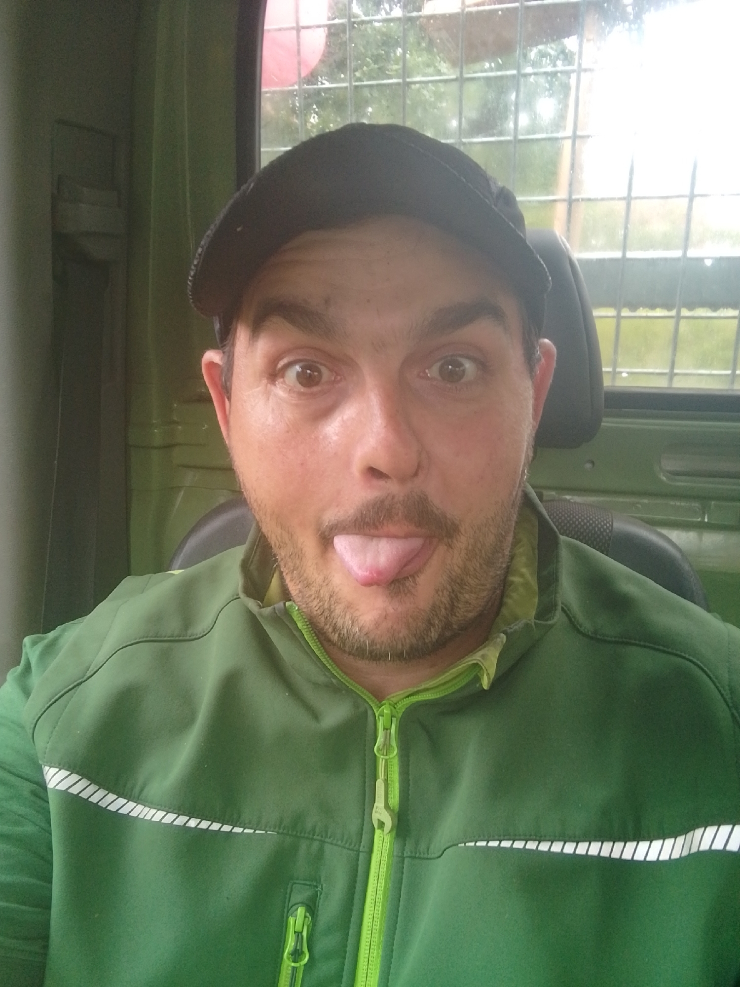A man is sitting inside a vehicle, wearing a green jacket and a black baseball cap. He is sticking his tongue out at the camera. The background appears to be the interior of the vehicle, with a window showing greenery and a grid-like structure. The lighting suggests it is daytime. There is no visible indication of the city of Hamburg, Germany.