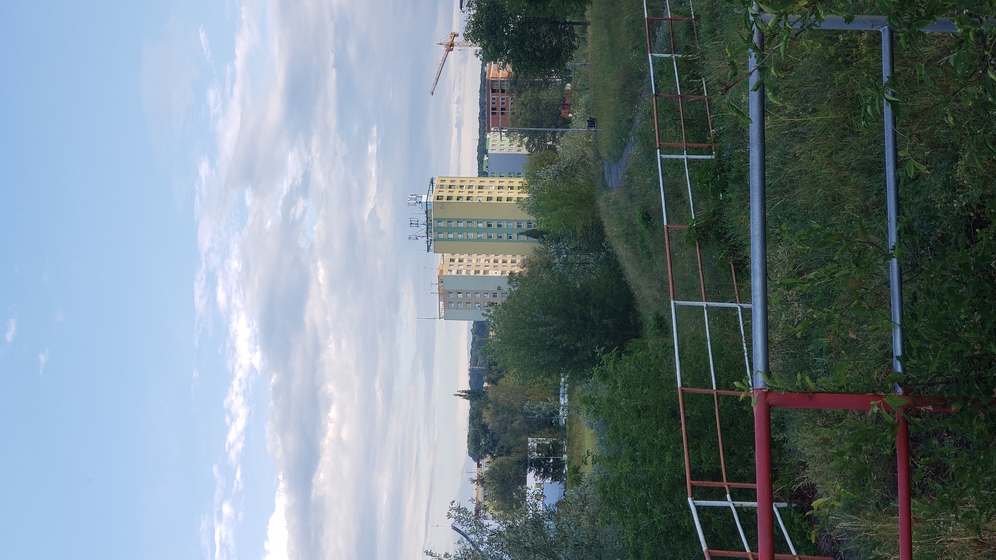 The image depicts a residential area with several tall apartment buildings under a cloudy sky. In the foreground, a red and silver metal railing separates a grassy area from what appears to be a path or embankment. The apartment buildings are multi-story structures with numerous windows. The tallest building in the center is light green with yellow accents and has antennas on its roof. To its left, a similar building is beige and grey. In the background, a construction crane is visible. Trees and greenery are scattered throughout the scene, suggesting an urban environment with parks or green spaces. The scene appears to be captured during daylight hours. This location is likely in Prešov, Slovakia, as the architecture and style of the buildings are characteristic of Eastern European residential developments.