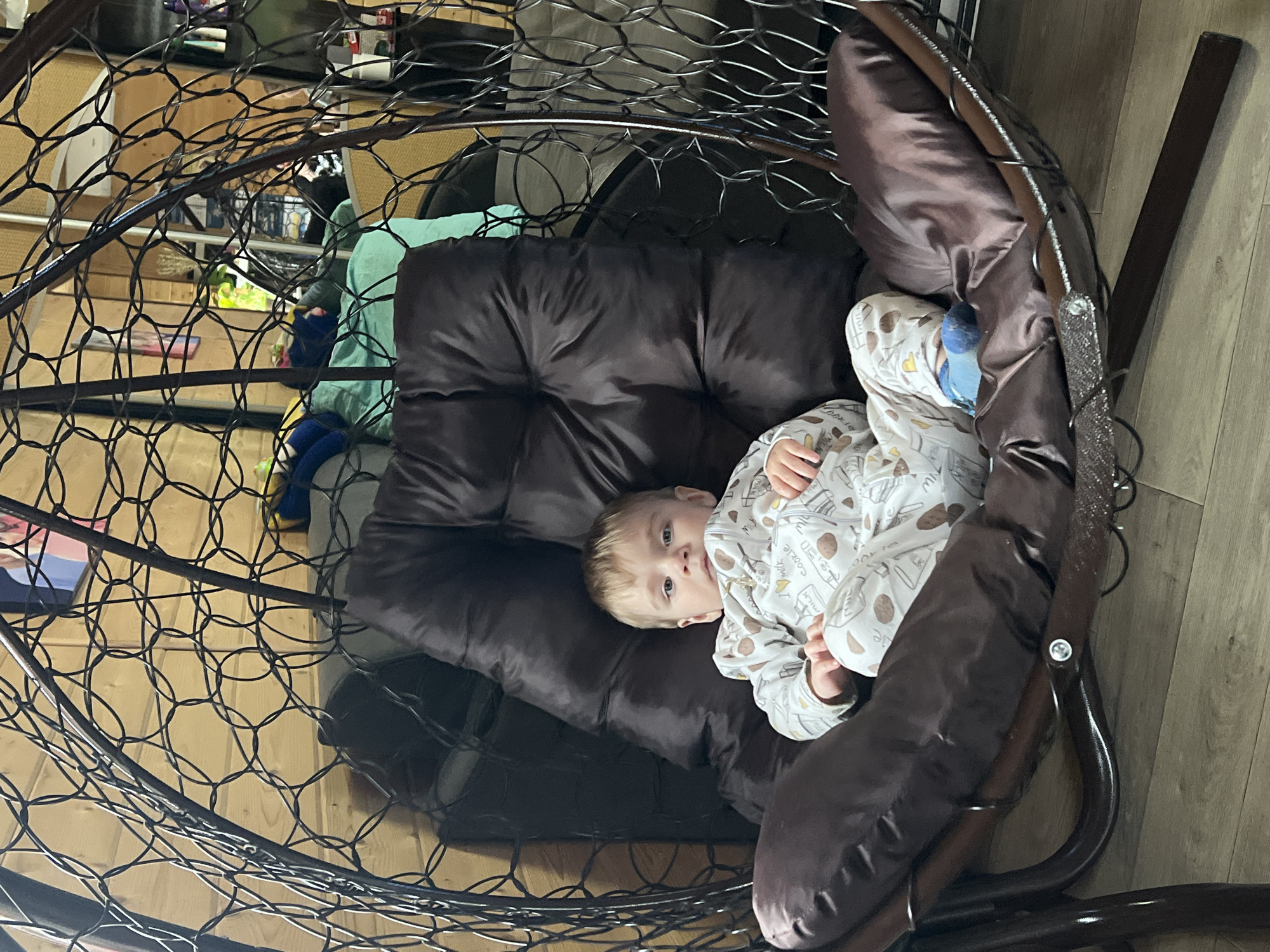 A baby is lying in a brown hanging chair indoors, likely in a home. The chair has a woven metal frame and a large, padded cushion. The baby is wearing a white patterned pajama suit and blue socks. The background shows a wooden floor and glimpses of items stored on shelves. The image was taken in the city of Protasy, Russia.