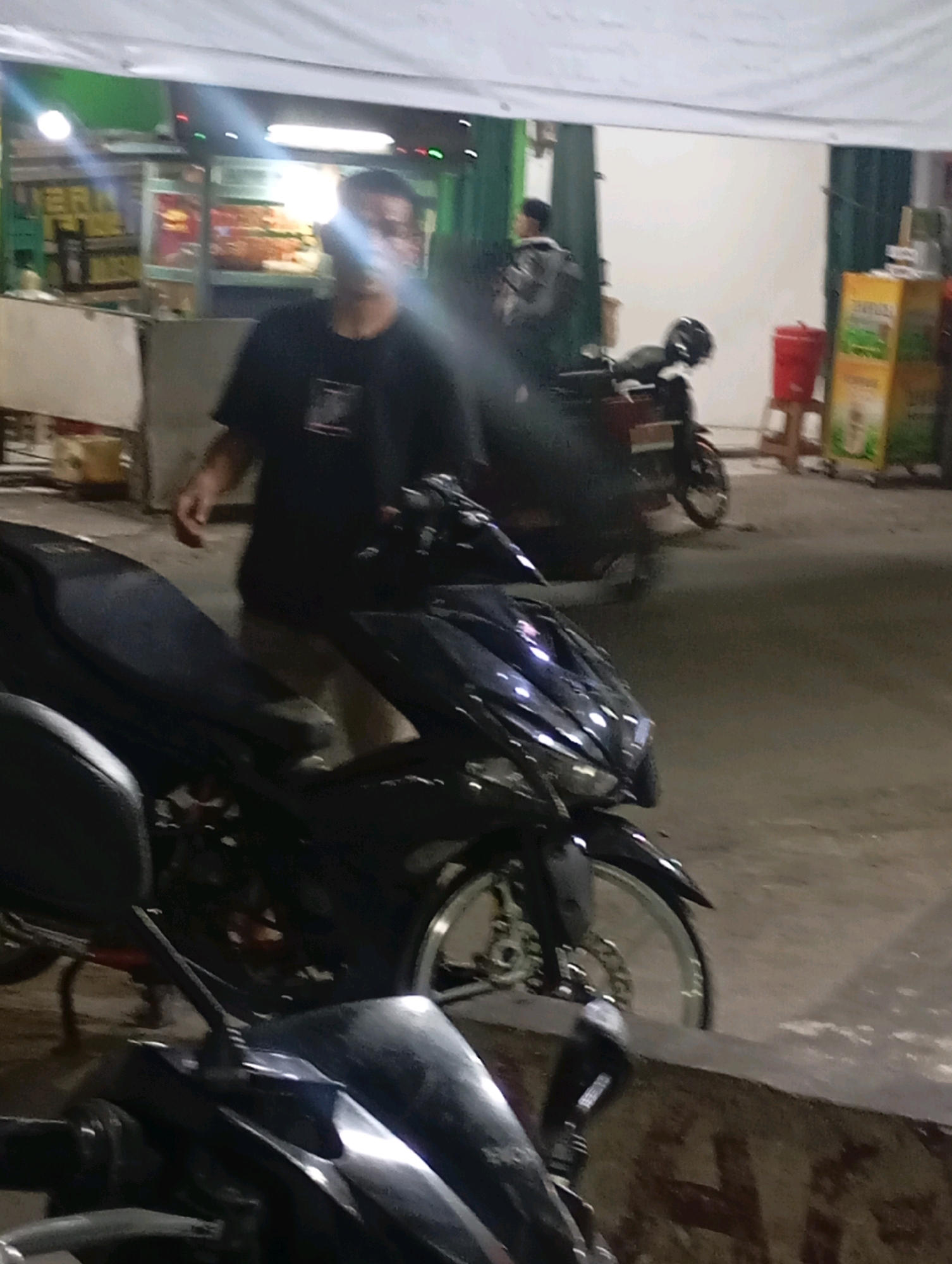 A blurry, low-light nighttime photograph taken at street level in Babelan, Indonesia, depicts a man in a black t-shirt and khaki pants standing next to a dark-colored scooter. The man is in the mid-ground of the image, partially obscuring the scooter. The scooter is parked on a paved surface, possibly a sidewalk or street, in front of a building with a green facade and a white awning. In the background, another person in a dark jacket is visible near a maroon scooter. To the right, a red plastic bin sits on a wooden stool next to a tall yellow sign with green lettering. The scene appears to be an outdoor food stall or market area, with a food stall structure visible to the far left. Lights are present, casting reflections and creating glare within the image.