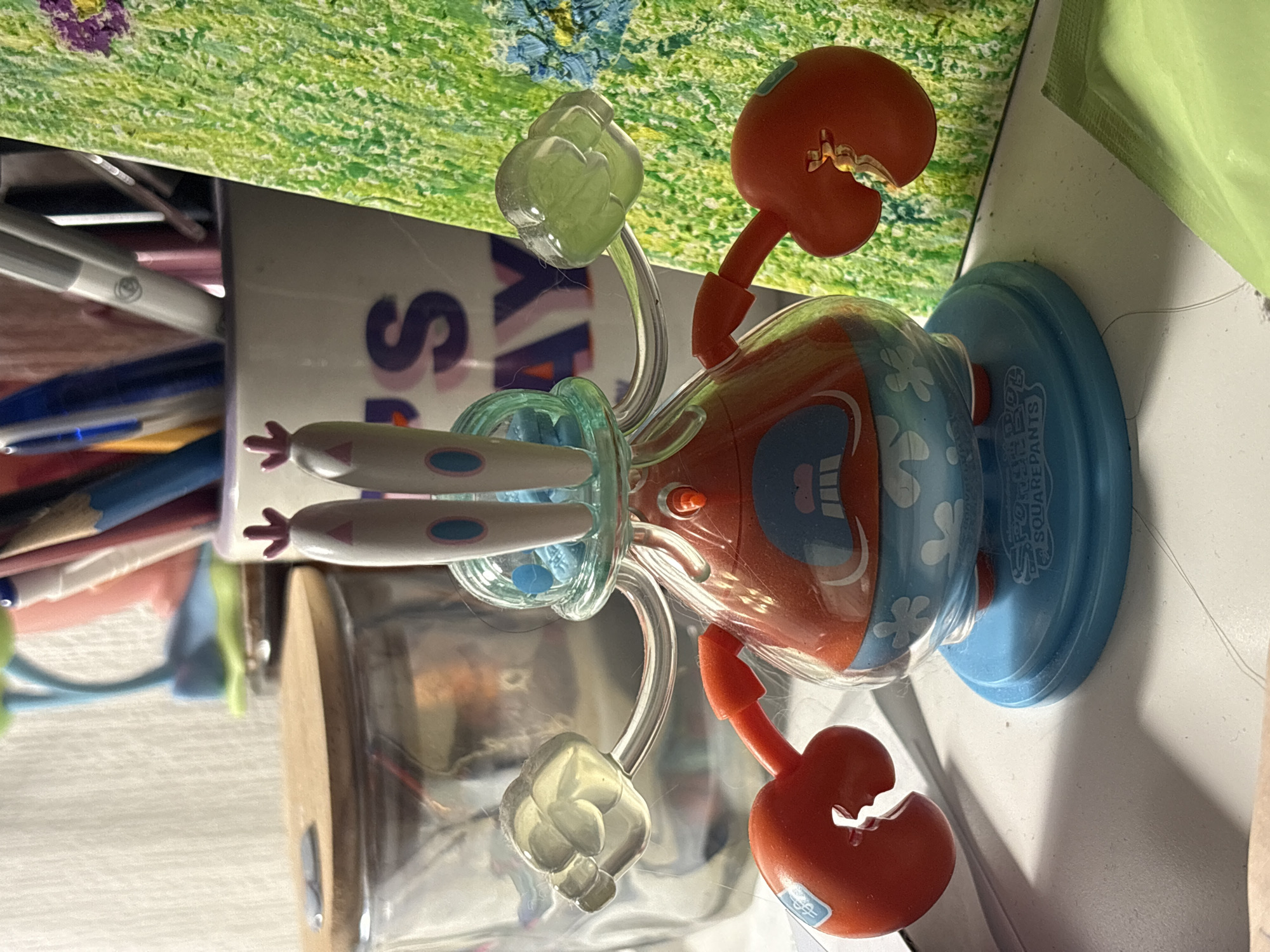 A Spongebob Squarepants toy, likely Mr. Krabs, stands on a blue base on a white desk. The toy has red crab claws, a translucent orange body with a blue and white patterned lower half, and a clear plastic dome on its head. Two clear, fist-shaped objects are attached to its sides, and two green, cloud-shaped objects are attached to thin silver arms that extend from the toy's body. The toy is positioned in front of a glass jar with a wooden lid and a colorful, textured background that appears to be a painting or drawing of grass. Several pens and pencils are visible in the background, along with a white mug with purple lettering. The overall scene suggests a desk or workspace. The location is unknown.