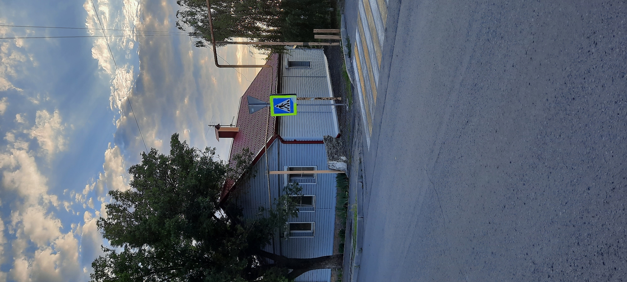 The image depicts a street scene in Shakhty City District, Russia, on a cloudy day with sunlight filtering through. In the foreground, a paved road with visible cracks occupies the lower half of the frame. A pedestrian crossing with yellow and white stripes is prominent on the right side of the road. Adjacent to the crossing, a blue and yellow pedestrian crossing sign is mounted on a post.
Behind the road and crossing is a light blue, single-story house with a red tiled roof. The house has several windows and a visible chimney. A large, mature tree with dark green foliage stands to the left of the house. The background features more trees and utility poles with wires. The sky is filled with a mix of white and gray clouds, with shafts of sunlight breaking through, creating a dramatic effect. The overall impression is a quiet, residential street bathed in the late afternoon or early morning light.