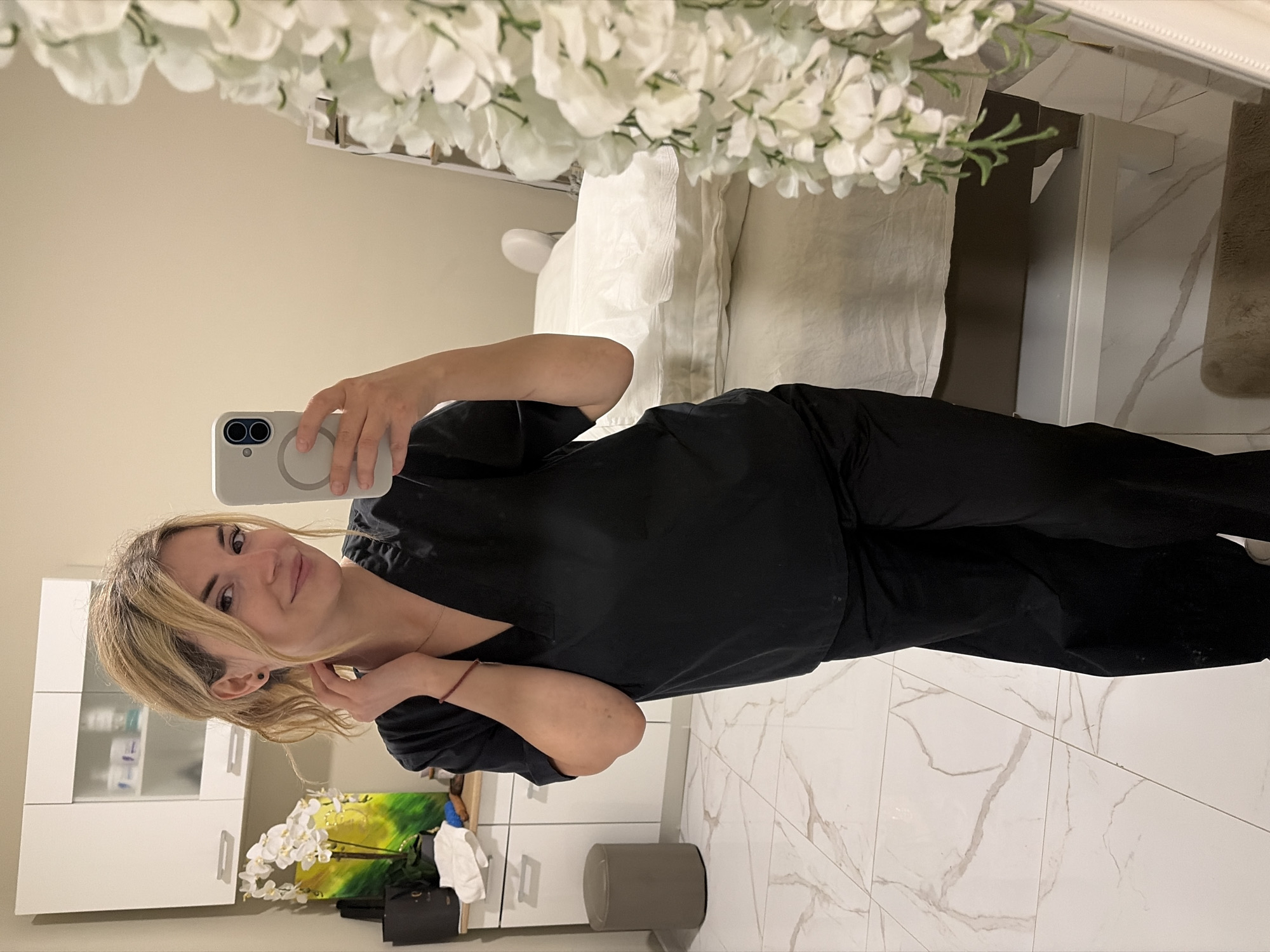 A blonde woman, wearing black scrubs, is captured taking a mirror selfie in what appears to be an aesthetic clinic or spa treatment room. She is holding a light grey smartphone with a MagSafe ring and dual cameras in her right hand, smiling gently at her reflection.
The setting is bright and modern, characterized by light beige walls and a white tiled floor with subtle grey veining. Directly behind the woman's reflected figure, a pristine white treatment bed or table, draped with white sheets, is visible. Above this bed, a reflection shows a decorative arrangement of abundant white artificial flowers with green accents, creating a canopy effect.
To the left side of the reflection, a sleek white cabinet with glass doors contains various bottles and products, suggesting a professional beauty or medical context. Below the cabinet, a decorative element features white flowers set against a vibrant yellow and green background, possibly a painting or another plant arrangement. A grey cylindrical stool or bin is placed on the floor. The overall ambiance is clean, professional, and inviting. No legible text is visible within the image. The location context for this scene is Budva, Montenegro.