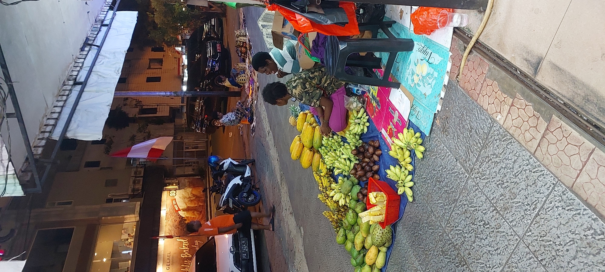 The image captures a street scene in Jayapura, Indonesia, likely at dusk or night. In the foreground, a vendor is selling a variety of fruits spread on a blue tarp laid on the ground. The fruits include papayas, mangoes, bananas, and what appear to be salak (snake fruit). Two women are seated near the fruits, one wearing a camouflage-patterned shirt and a purple skirt, and the other in a striped green and white shirt. A black plastic stool is placed beside them. In the mid-ground, a man in an orange t-shirt and black shorts stands near a white motorcycle. Behind him, a white car is parked. Further back, several parked cars are visible, including dark SUVs, and a building with illuminated windows. A prominent Indonesian flag, red and white, is flying from a pole. The ground is paved with cobblestone-like tiles in the foreground and grey textured tiles further back. The scene suggests a typical roadside market activity in the city.