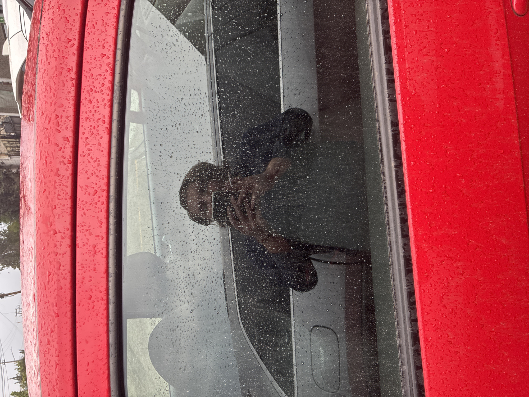 The image shows the exterior of a red vehicle, possibly a bus or truck, with a window reflecting a person taking a photo with a mobile phone. Raindrops are visible on the window and the red exterior. The reflection suggests the interior is dark, with grey seats. The reflection shows a person holding a phone, taking a selfie. The outside is a rainy day. It is not possible to determine the location in Cuautitlán Izcalli, Mexico, based on the provided image.