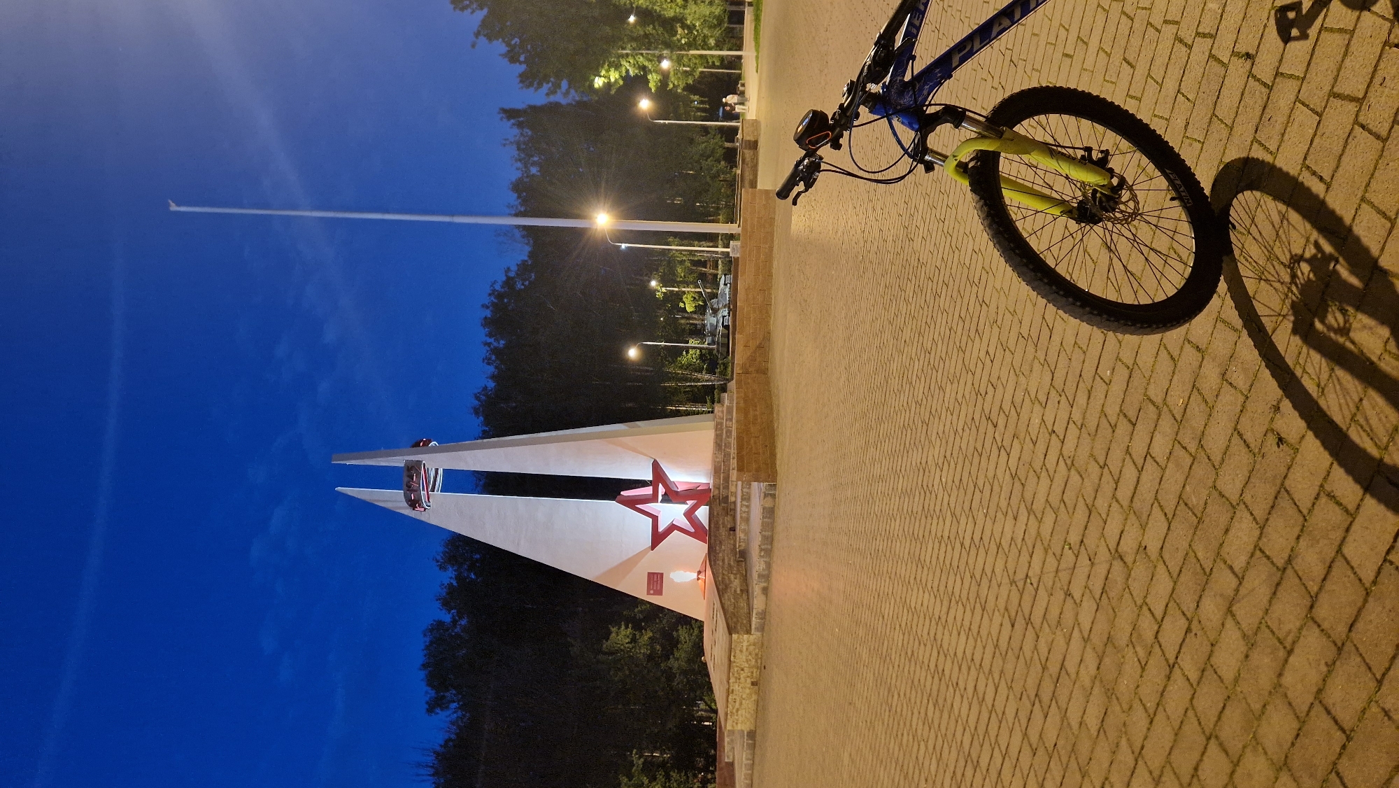 In the city of Otradny, Russia, at dusk, a bicycle is parked on a paved plaza in front of a large, white, V-shaped monument. The monument is illuminated and features a prominent red star at its base, with an eternal flame burning to the left of the star. Above the star, a circular element with red and blue accents is visible at the apex of the monument.
In the background, a dense line of trees forms a dark backdrop against the deep blue twilight sky. Several streetlights are visible, casting a warm glow on the plaza and the surrounding trees. To the right of the monument, a military tank is displayed, suggesting a memorial or historical significance of the location.
The bicycle in the foreground is blue and yellow and is partially obscuring the paved area, which is made of brick-like pavers. The bicycle's shadow is cast long across the ground. The overall scene conveys a sense of quiet solemnity, perhaps a memorial park or a public square dedicated to historical events.