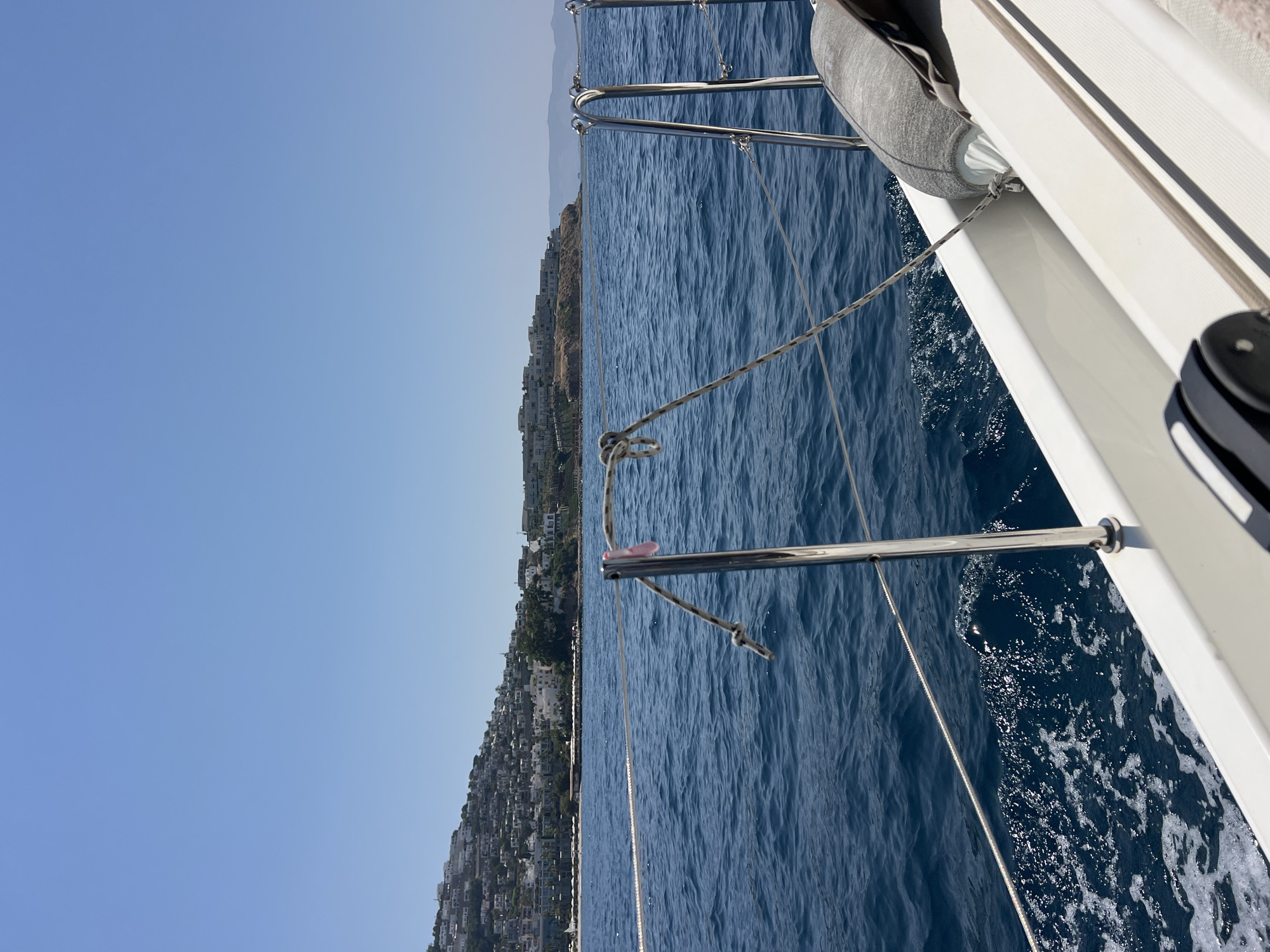 A forward-facing, wide shot captures a view from a sailboat moving through deep blue water under a clear, cloudless sky. The sailboat's white hull and chrome railings are visible in the foreground, with a gray fender secured to the railing. White foamy wakes trail behind the boat, indicating movement.
In the middle ground, the boat is sailing towards a coastline dotted with numerous white buildings constructed on a steep hillside. The buildings are densely packed and appear to be residential, with some structures reaching higher up the hill. The coastline stretches across the horizon, meeting the water with a thin strip of what appears to be a beach or shoreline.
In the background, the horizon is marked by hazy, faint mountain ranges under the expansive blue sky. The overall scene suggests a sunny day on the water, with a coastal town visible in the distance.