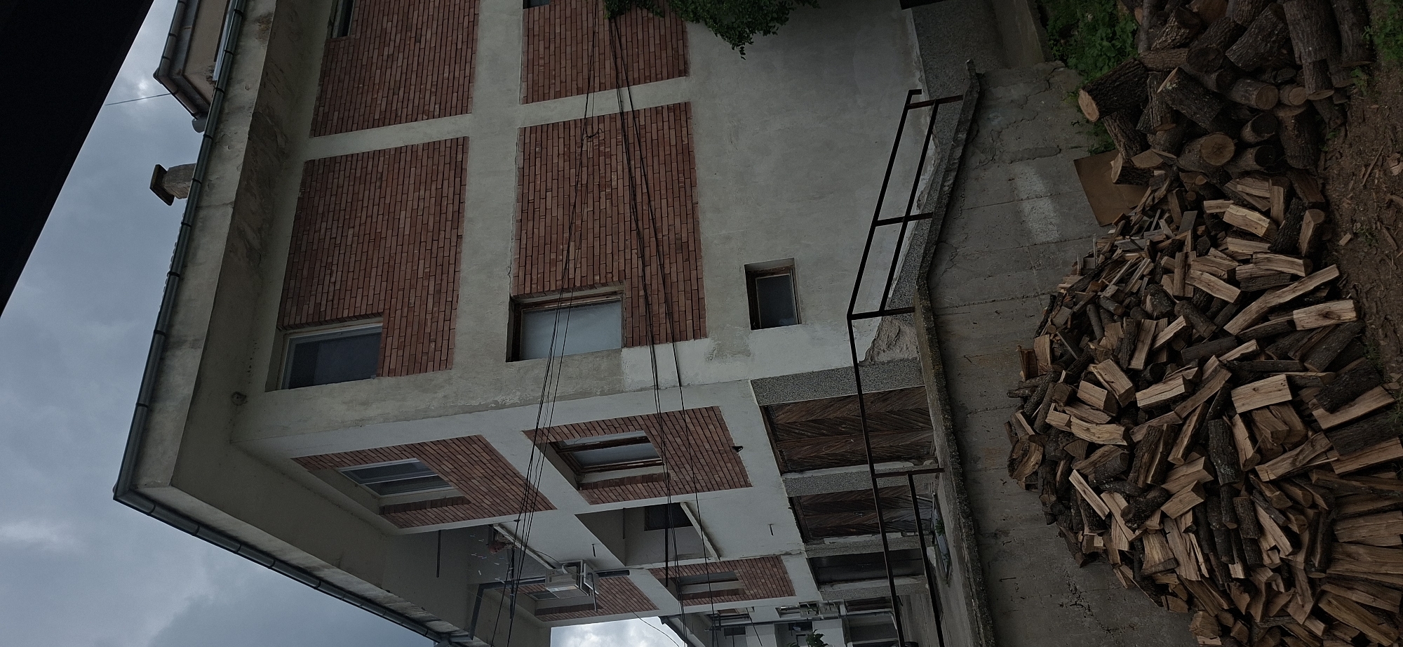 The image shows an outdoor scene, rotated 90 degrees clockwise from its natural upright orientation.
The main subject is a multi-story building facade, which occupies the left and central-left portion of the frame. The building features a light grey, possibly concrete or stucco, exterior interspersed with large, rectangular panels of red brickwork arranged in a grid pattern. Several windows are visible; some reflect the grey sky, while others appear as dark openings. Dark rain gutters are present along the visible edges of the building's rooflines. Black cables or wires hang vertically down the building's side. Towards the bottom, an overhanging section of the building contains further windows or skylights, also framed by brickwork.
To the right of the building, a substantial pile of chopped firewood, consisting of logs and smaller pieces, is stacked on an uneven surface of concrete and dirt. A black metal, ladder-like structure stands on the concrete ground, extending upwards towards the building. Some green foliage is visible in the upper right corner of the frame.
The sky, visible towards the top-left, is uniformly grey and cloudy, indicating overcast weather conditions. The setting appears to be an exterior utility area or backyard of a building, located in Strazhitsa, Bulgaria. No people or visible text are present in the image. The scene is static, capturing a functional outdoor environment.