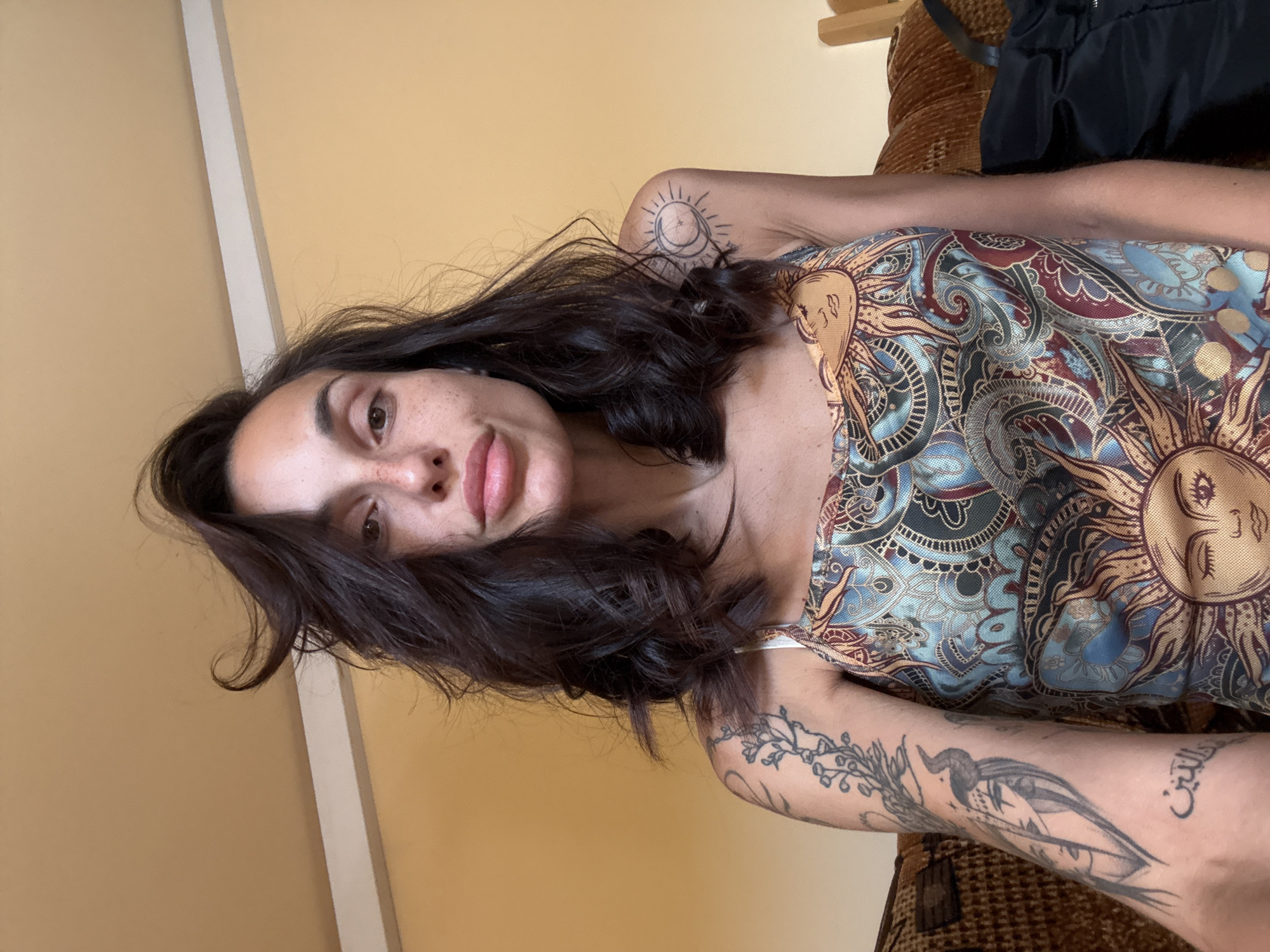 A woman with dark, wavy hair and freckles across her nose and cheeks is shown from the chest up, looking directly at the camera. She is wearing a sleeveless top with a colorful, ornate pattern featuring suns and swirling motifs. She has several tattoos visible on her left arm, including an image of a woman with horns and another with Arabic script, and a tattoo of a sun on her right shoulder. The background is a plain, light yellow wall with a white trim near the ceiling. She is seated on a brown, textured couch. The overall setting appears to be indoors, likely a domestic environment.