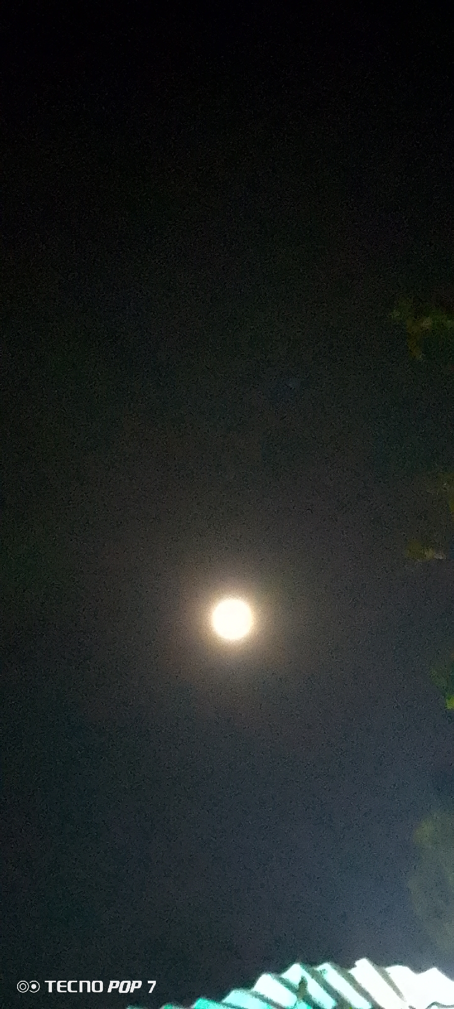 At night, the moon shines brightly in the dark sky over Altagracia, Venezuela. Partial rooftops and green foliage are visible along the bottom and right sides of the frame. The "Tecno Pop 7" watermark is in the lower left corner.
