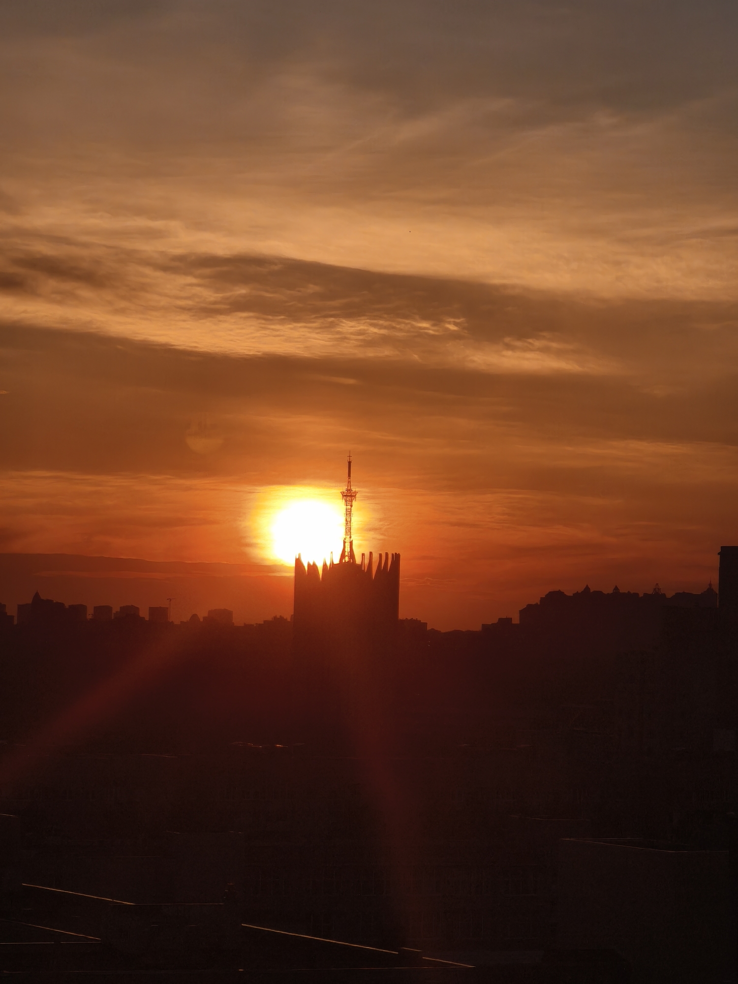The image depicts a sunset over Saint Petersburg, Russia. The sun is positioned behind a silhouette of buildings, including the Peter and Paul Cathedral, notable for its tall spire topped with a golden angel. The sky is a mix of orange and red hues. The foreground is obscured in shadows, possibly showing details of building tops. There are no people visible in the scene.