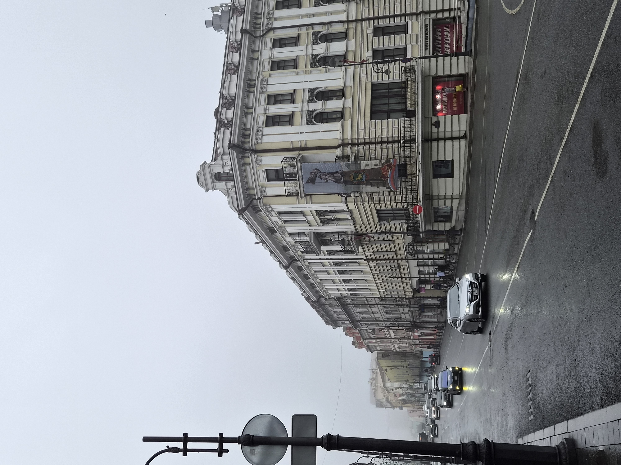 A street scene in Vladivostok, Russia, on a foggy day. The road is wet, suggesting recent rain, and reflects the light from car headlights and street lamps. Several cars are driving on the road, some with their headlights on. On the right side of the street is a multi-story building with ornate architecture, painted in light colors with decorative elements. A large banner with imagery of a statue and red stars is hanging from the building. The ground floor of the building appears to be commercial, with signs in Russian. On the left side of the street, a lamppost with traffic signs is visible, as well as more buildings in the background fading into the fog. A few pedestrians are visible on the sidewalk near the commercial establishments. The overall atmosphere is subdued due to the weather and lighting conditions.