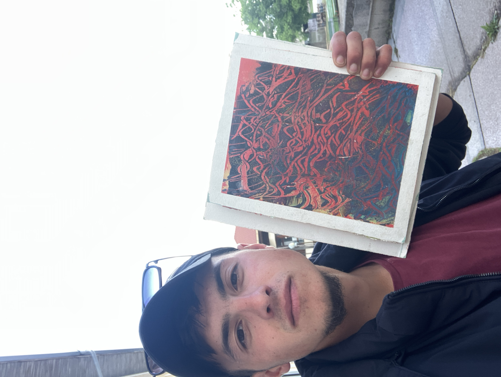 A man is holding up a stack of artwork in an outdoor setting. The artwork has a red and black abstract pattern on a white background. The man is wearing a black jacket over a maroon shirt and a black baseball cap. Sunglasses are visible hanging from his shirt. The background appears to be an urban environment with a hint of greenery, likely Krastevich, Bulgaria.