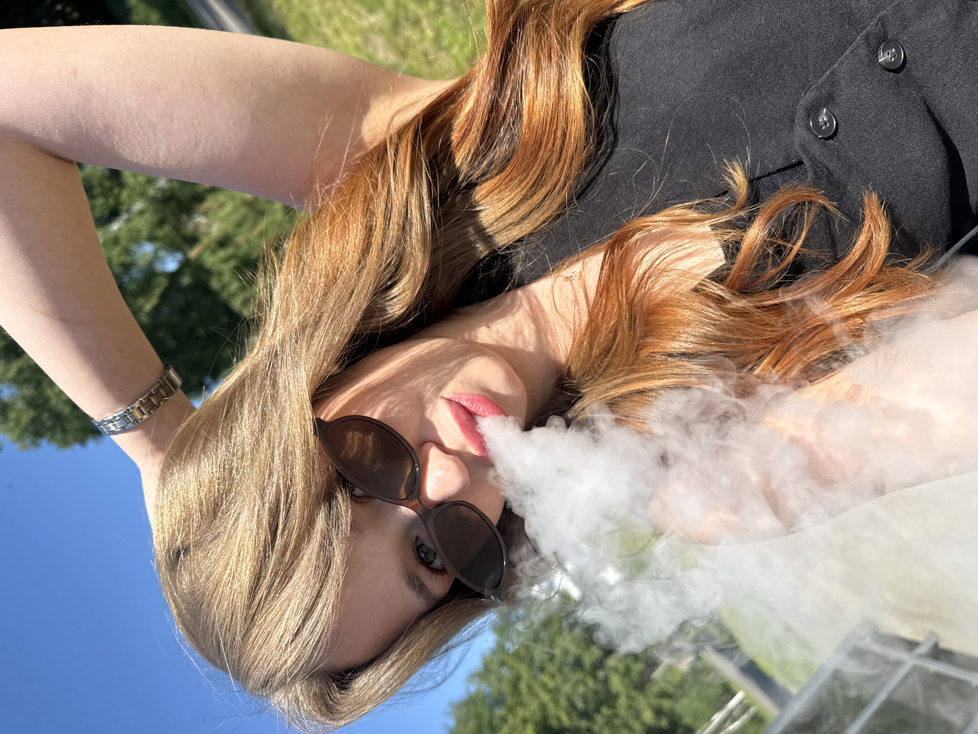 A young woman with long, wavy blonde and reddish-brown hair is captured in a close-up shot, outdoors under a bright blue sky. She is wearing sunglasses and a black vest. Her right arm is raised, with her hand behind her head, and she is exhaling a plume of white vapor. The background shows blurred greenery, suggesting a park or natural area. The image does not provide any specific indicators of the city of Chaikovsky, Russia.