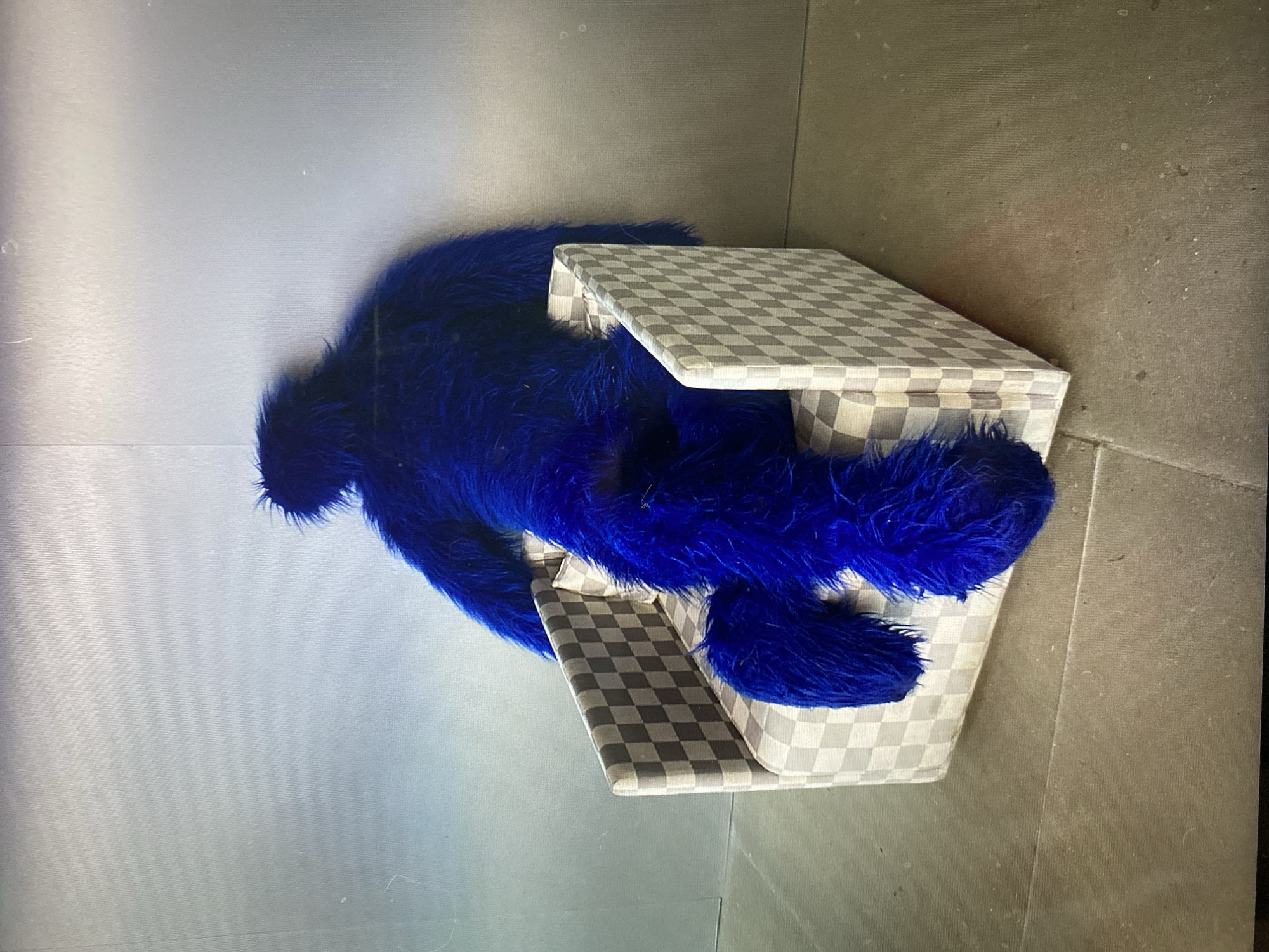 A fluffy, bright blue creature, appearing to be a stuffed animal or costume, is seated backward in a small, checkerboard-patterned armchair. The creature's large, bushy limbs are draped over the sides and front of the chair. The armchair itself is covered in a gray and white checkerboard fabric and sits on a grey, tiled floor. The background is a plain, metallic-toned wall. There is no discernible indication of the location within the city of Khimki, Russia.