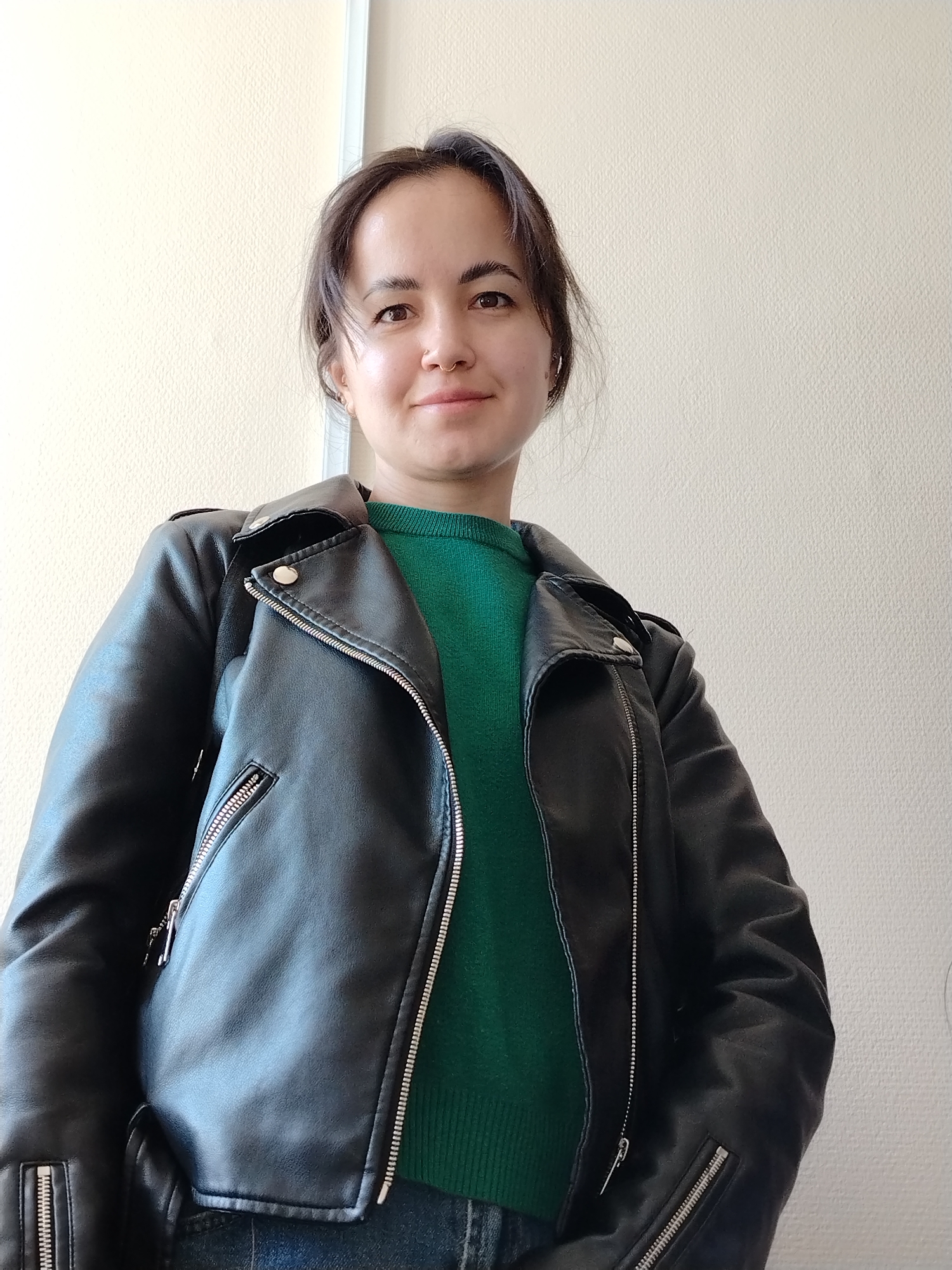 A woman stands facing the camera. She has dark hair pulled back from her face, brown eyes, and light skin. She is wearing a green sweater and a black leather jacket, which is partially unzipped. The background is a plain, light-colored wall with a vertical white strip on the left side. The location is likely indoors. It is impossible to determine if this image was taken in Chaikovsky, Russia with the context provided.