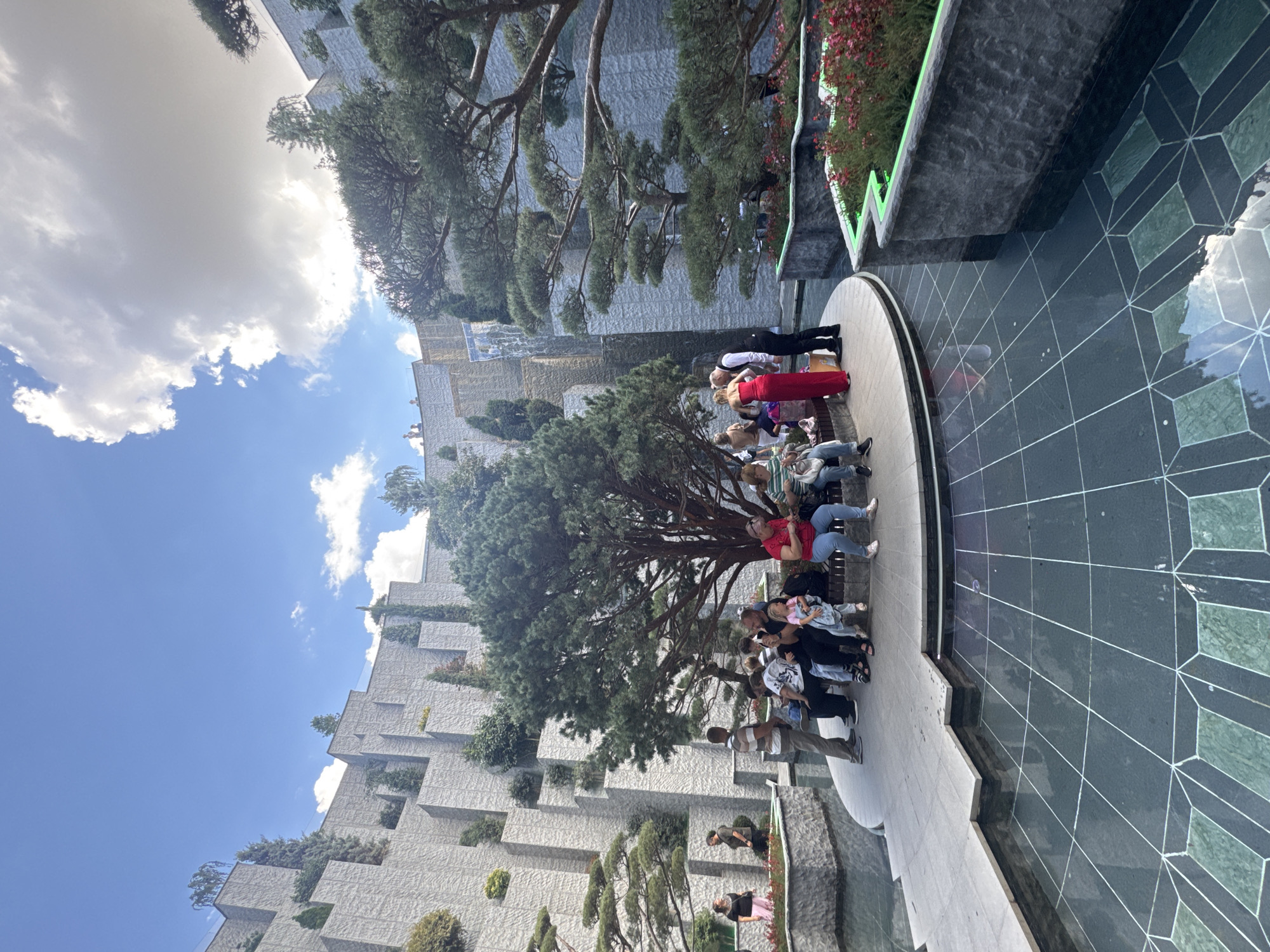 The image depicts a large, multi-tiered water feature in an urban park setting. The architecture is characterized by angular, light-colored stone walls that form terraces and cascades, with lush green trees and smaller plants integrated into the design. A prominent pine tree with sprawling branches sits centrally in the foreground, shading a group of people seated on a curved bench. Other individuals are standing and walking around the water basin, which has a patterned tiled bottom. In the background, a waterfall flows down one of the stone structures. The sky above is partly cloudy. This location is identified as the "Old Park" in Krasnodar, Russia.